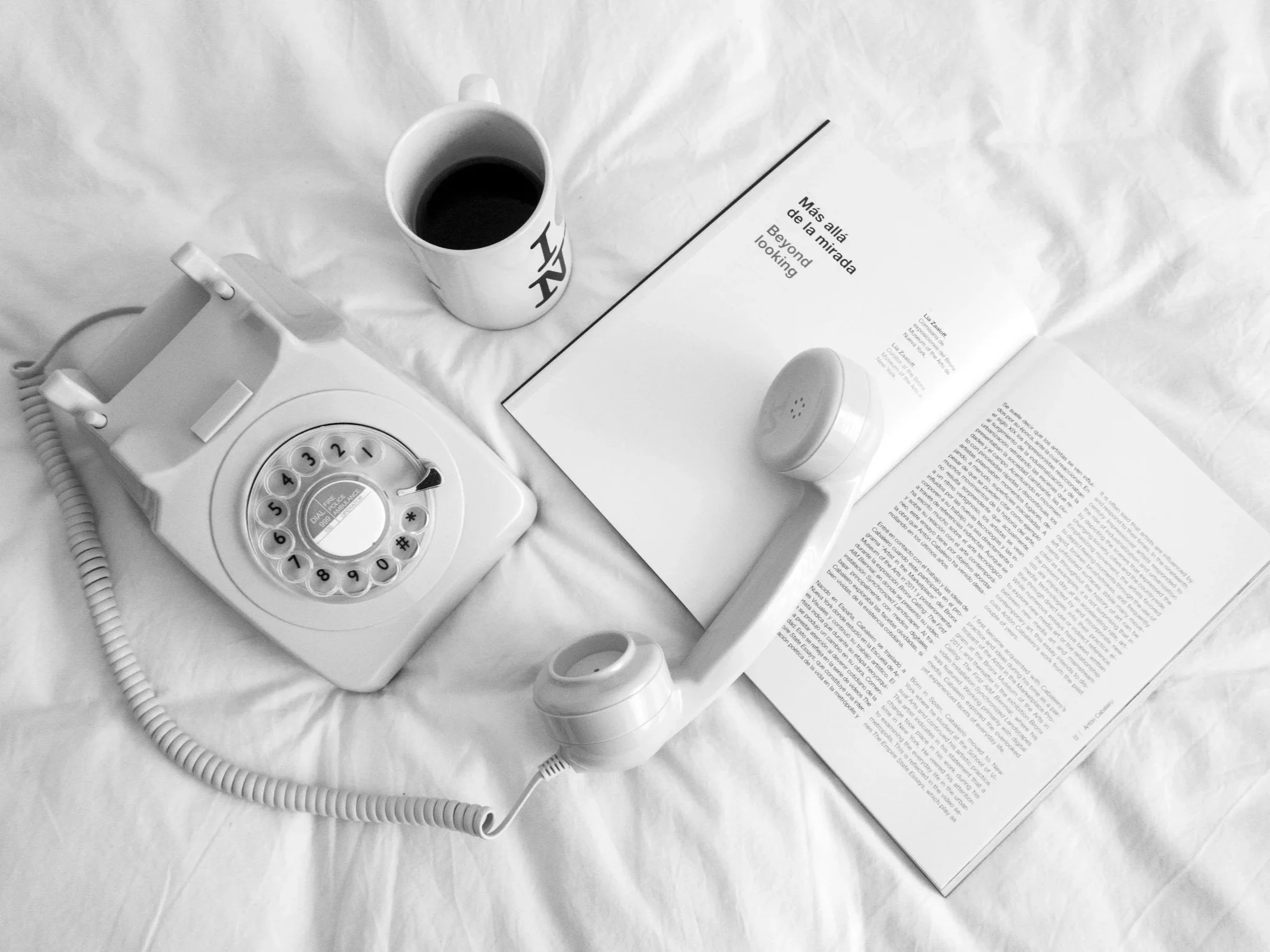 A flat lay of a white rotary telephone, an open book with text in Spanish and English, a cup of black coffee, a small cylindrical speaker, and an empty mug on a crinkled white fabric surface.