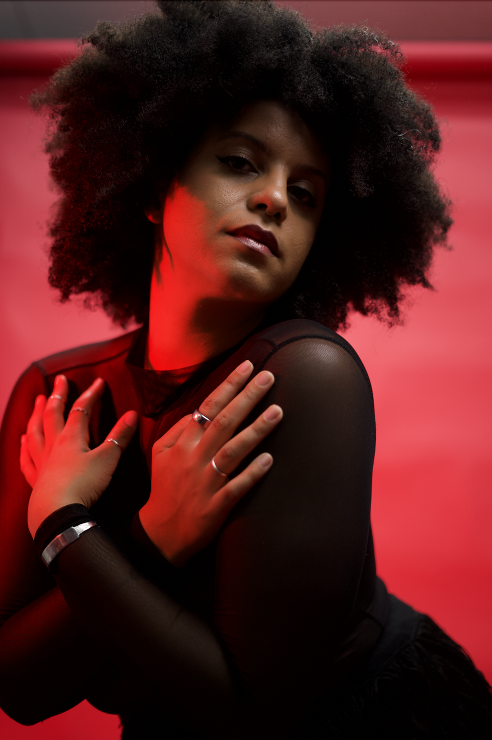 A woman with voluminous curly hair posing with her hands crossed over her chest, wearing black sheer and mesh clothing, illuminated by red and pink lighting.