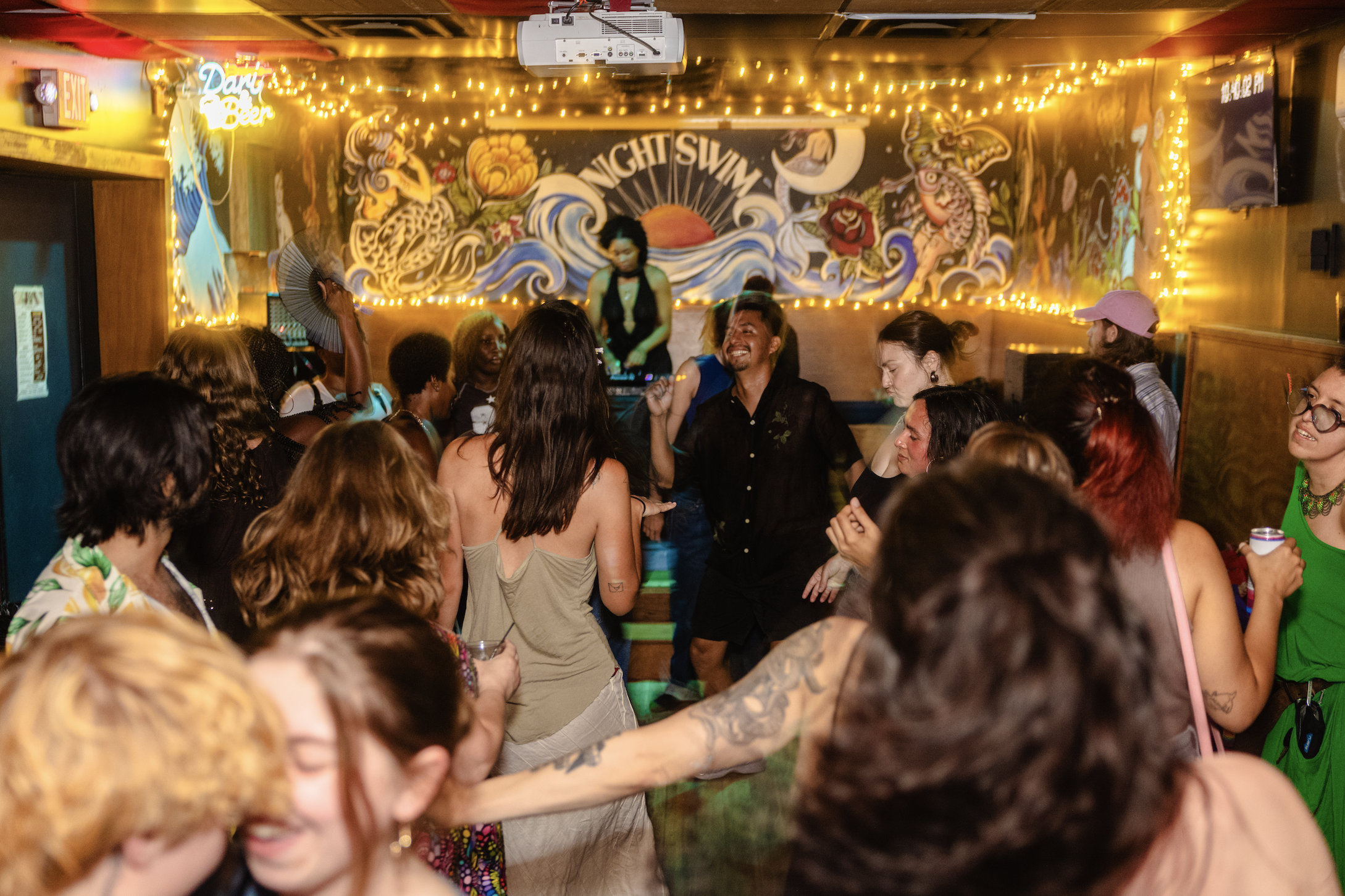 Crowd dancing at a lively indoor party with colorful wall art, string lights, and a DJ in the background.