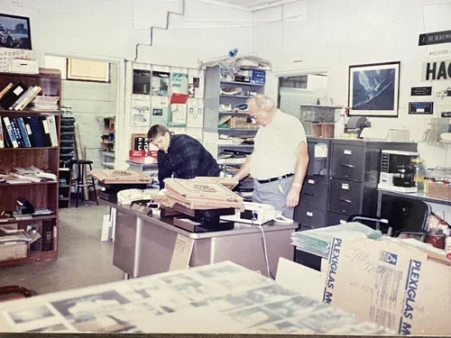 Two people working at a cluttered office desk surrounded by stacks of newspapers and files, with shelves filled with books, binders, and office equipment in the background.