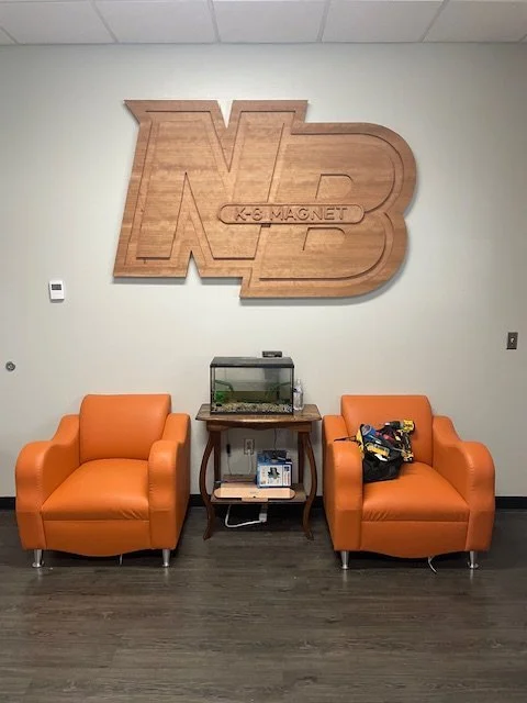 Office lobby with two orange armchairs, a fish tank on a wooden table, and a large wooden N3 logo on the wall with 'K-8 Magnet' engraved.