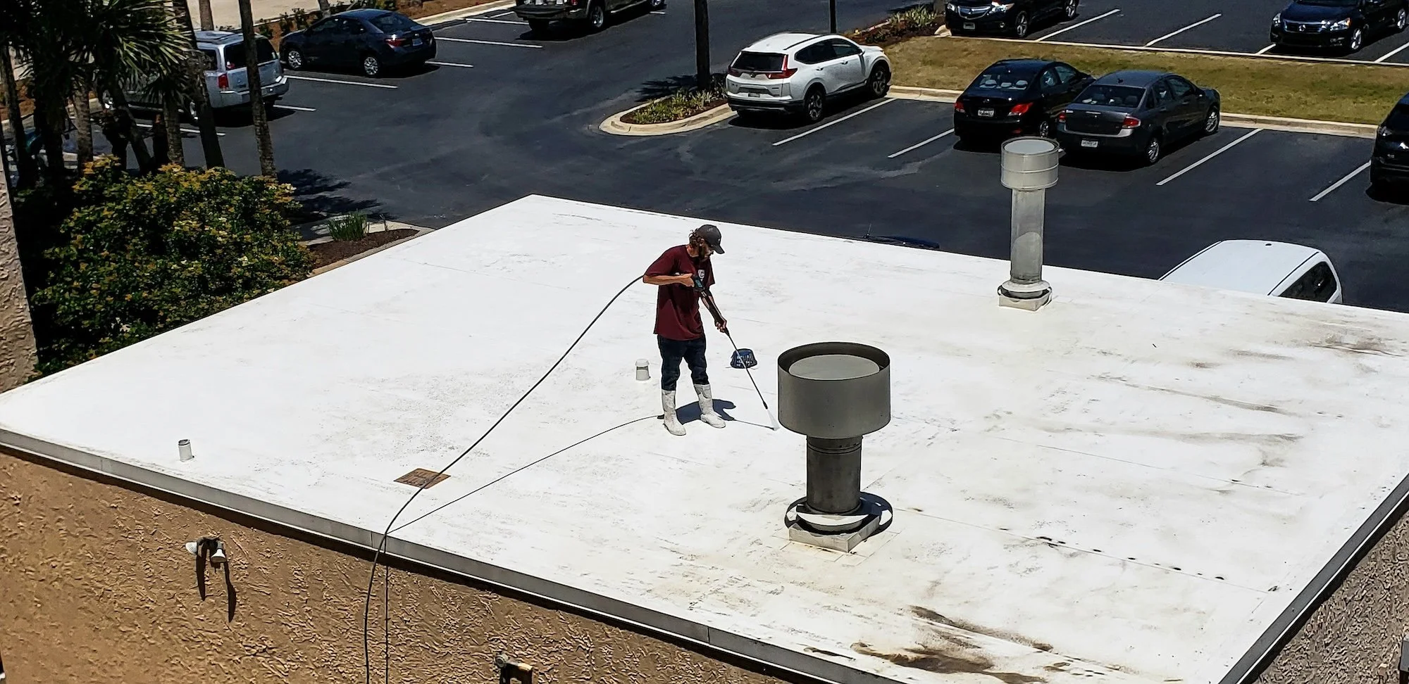 A person wearing white boots, a maroon shirt, and a cap is pressure washing the flat white roof of a building. The roof has several vents and a chimney. In the background, there is a parking lot with cars and a grassy area with trees.