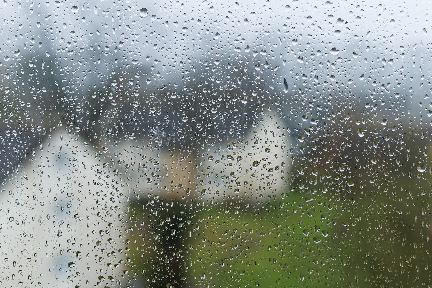 Rain droplets on a window with blurred houses and trees in the background.