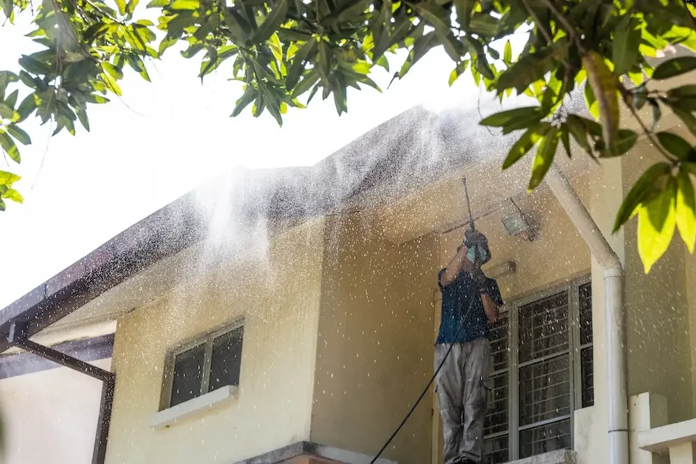 Man pressure washing the exterior of a beige house on a sunny day, seen through green leaves.
