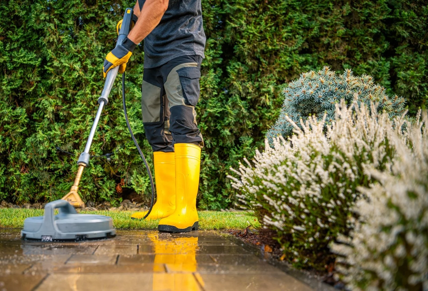 Person wearing yellow rain boots and waterproof pants using a pressure washer to clean a stone walkway outdoors, with green bushes and plants in the background.