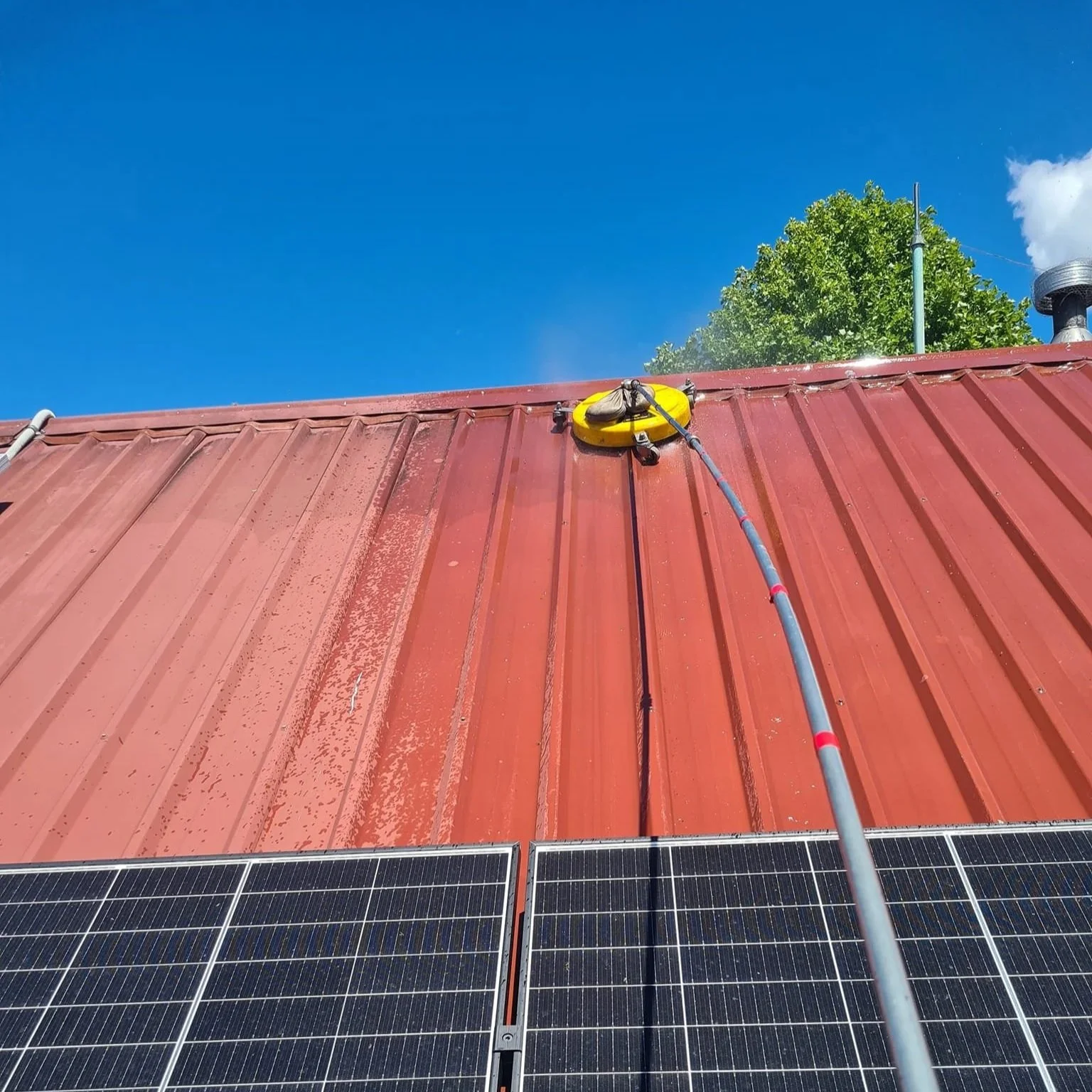 A solar panel installed on a red metal rooftop with a cleaning device attached, under a clear blue sky with some clouds and a green tree in the background.
