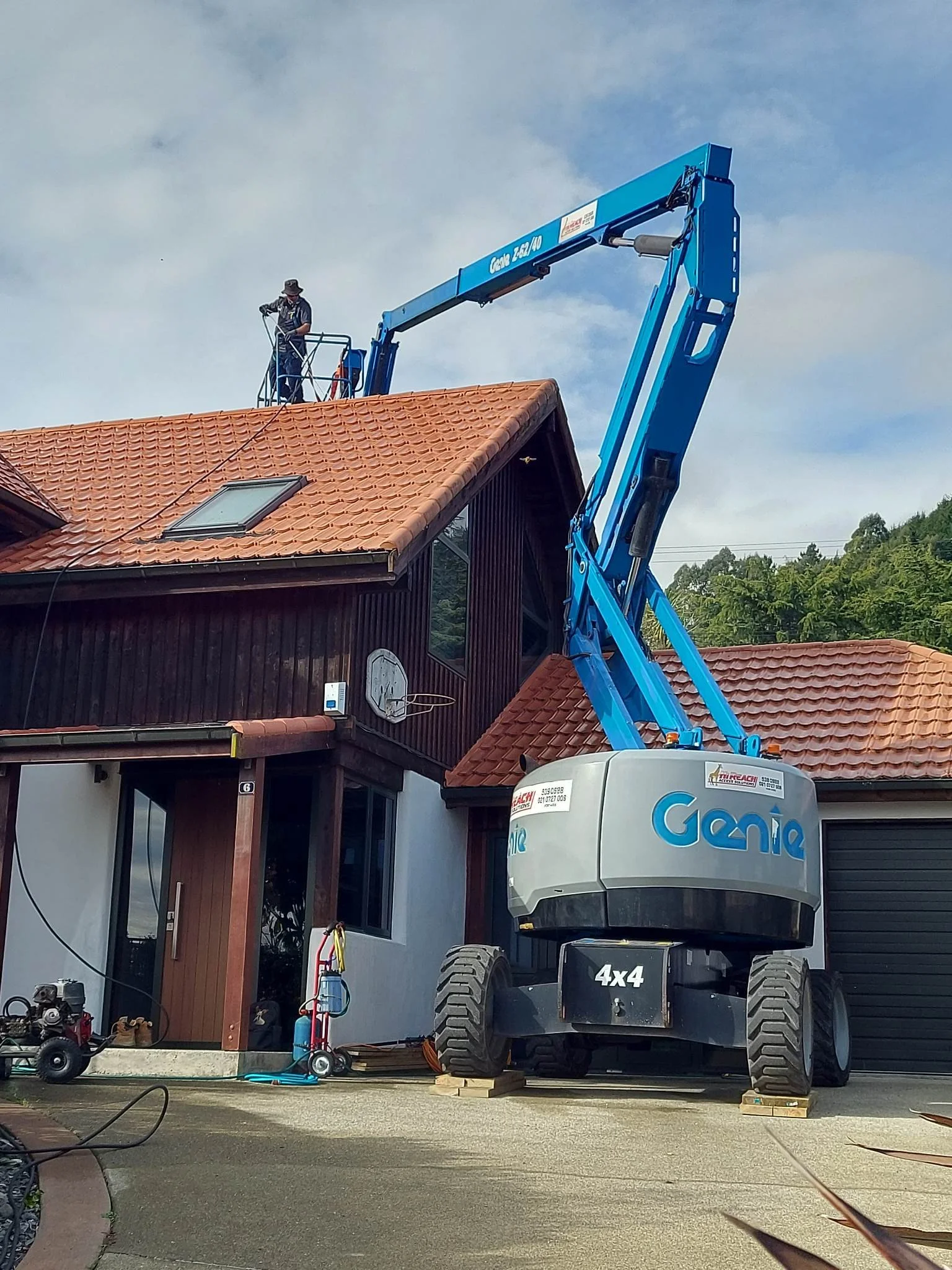 A person wearing a hat working on a blue lift platform on the roof of a house. The house has a red-tiled roof, and there are tools and equipment nearby.
