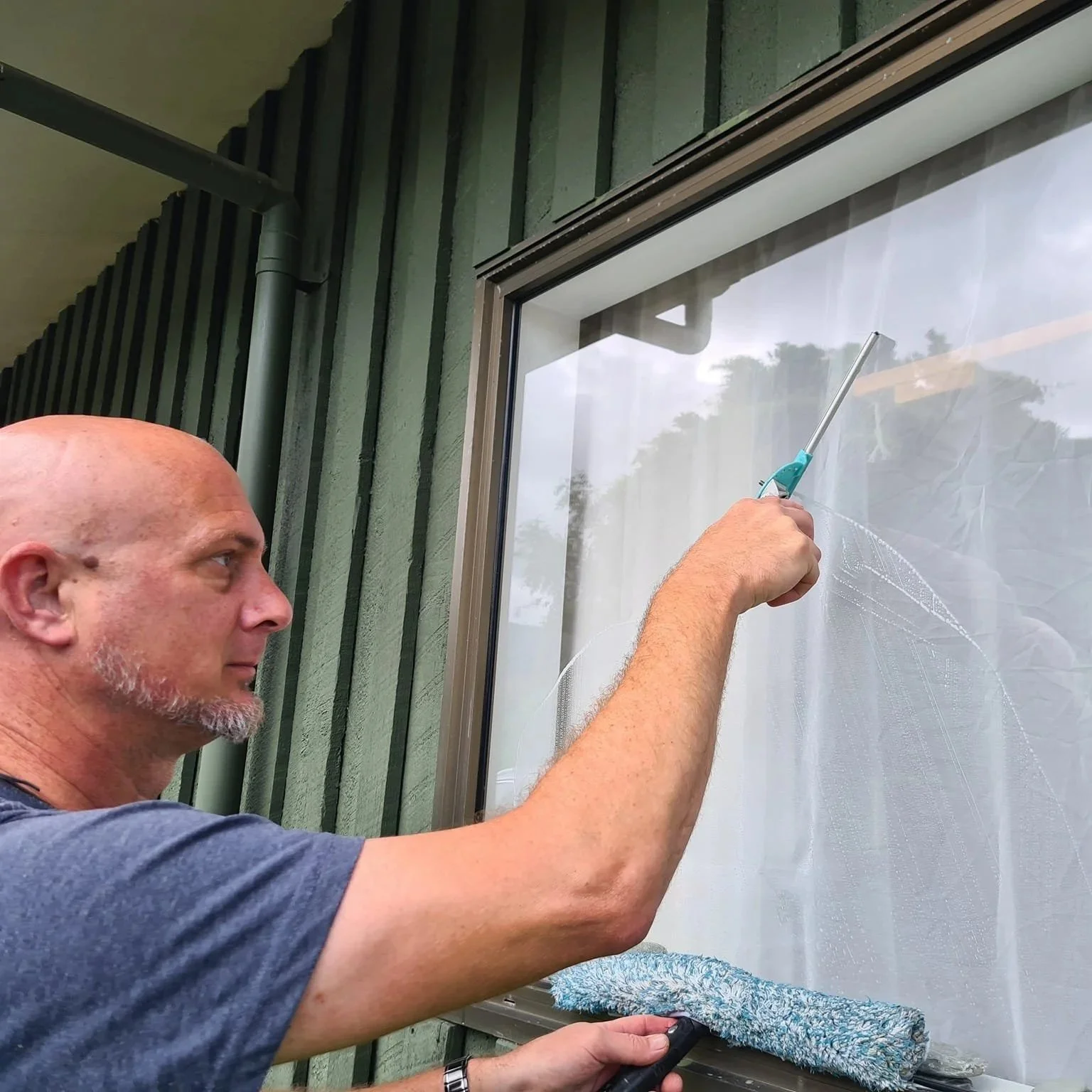 A man cleaning a window with a spray bottle and a cleaning roller, standing outside a green building.
