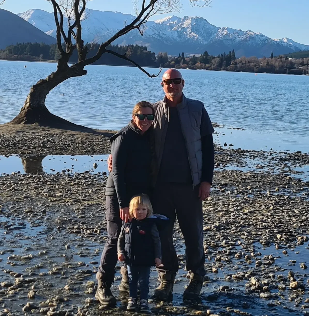 A family of three stands on a rocky lakeshore with a large, twisted tree in the background, snow-capped mountains, and clear blue sky in the distance.