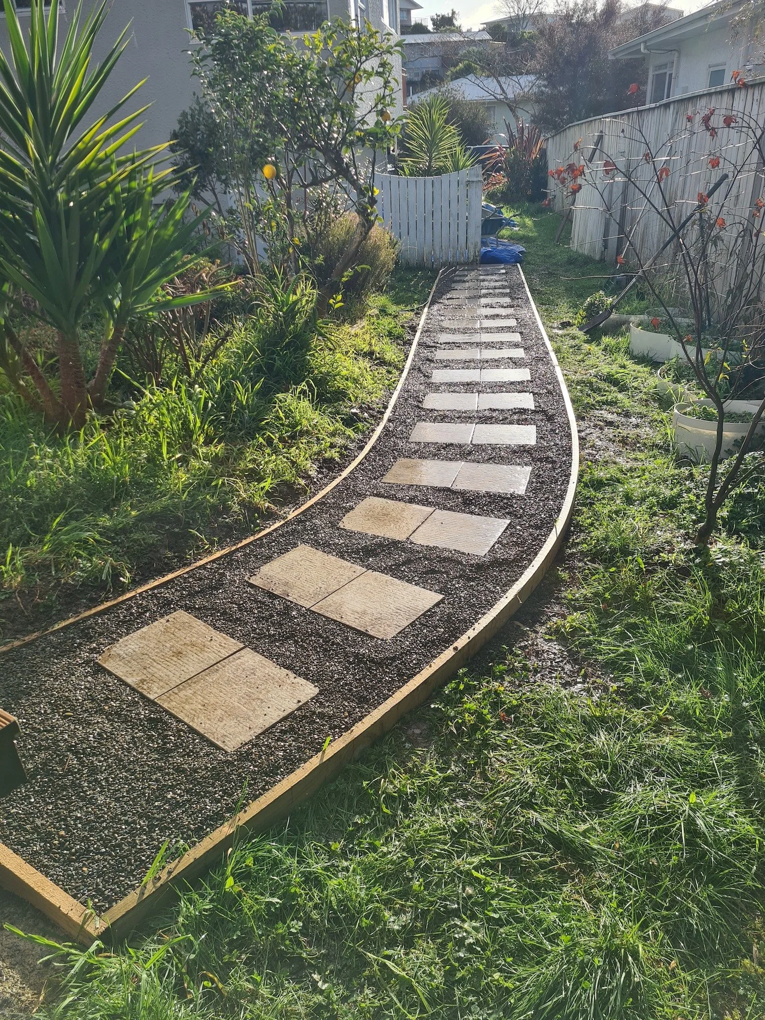 A garden pathway made of rectangular pavers embedded in a gravel base, flanked by lush green grass and various plants, leading to a white gate with a backyard beyond.