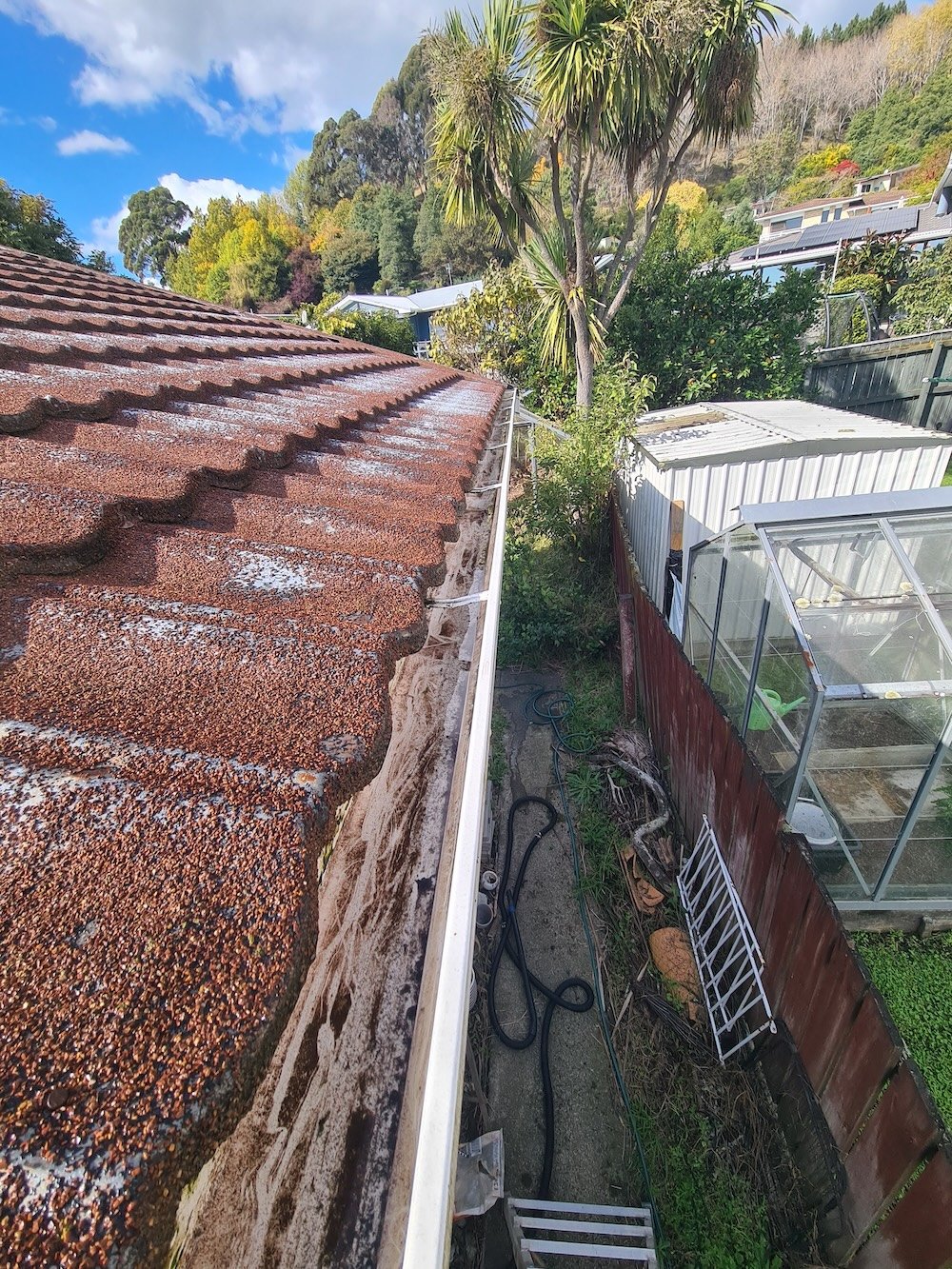 View from a rooftop looking down a narrow alleyway between houses. The roof has weathered, reddish-brown shingles. On the right, a red wooden fence borders a garden with a glass greenhouse. Behind the greenhouse, there are trees, shrubs, and neighboring houses on a hillside. The sky is partly cloudy with blue patches.