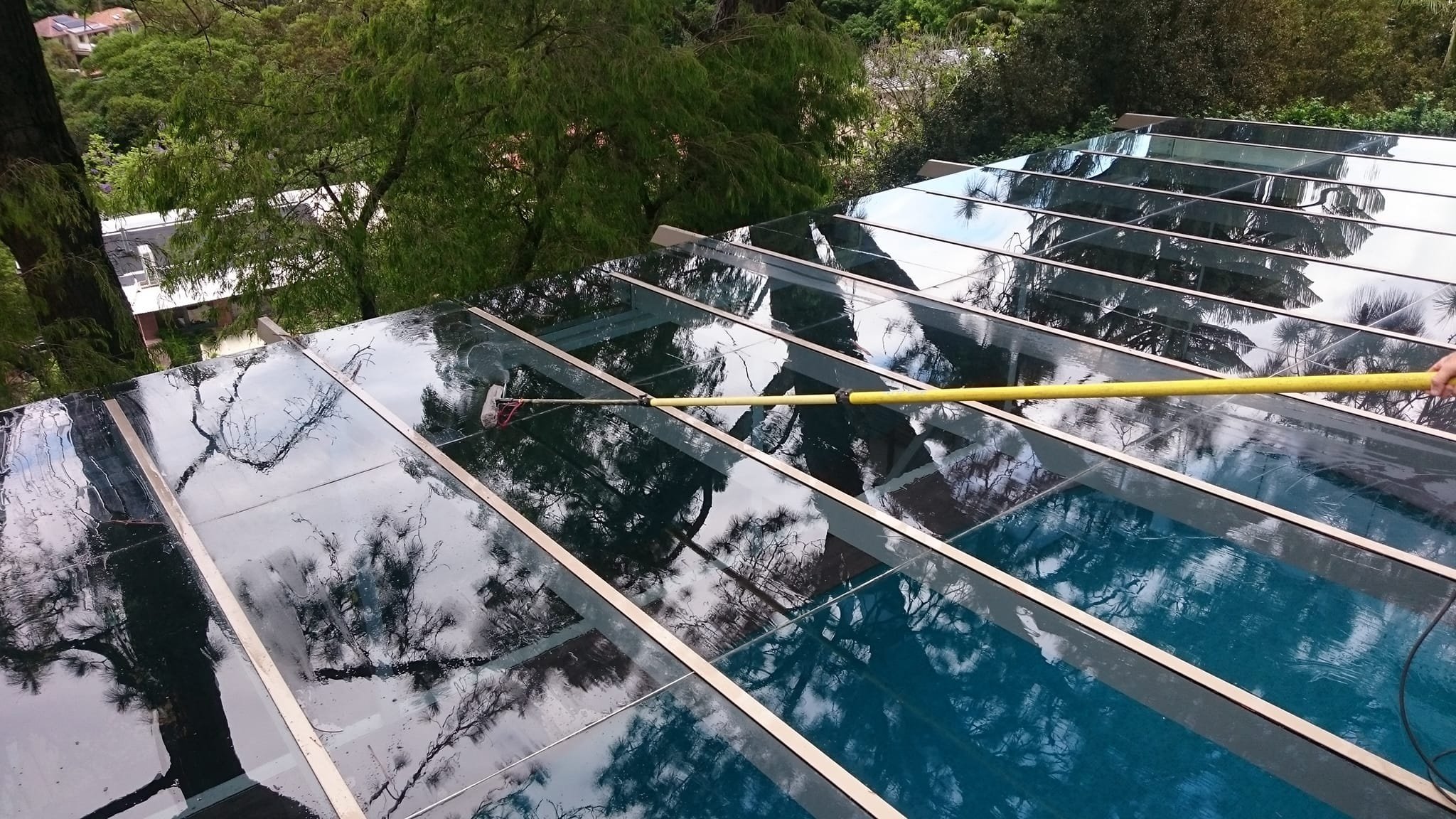 Solar panels on a roof reflecting the sky and surrounding trees, with a person cleaning the panels using a yellow tool.