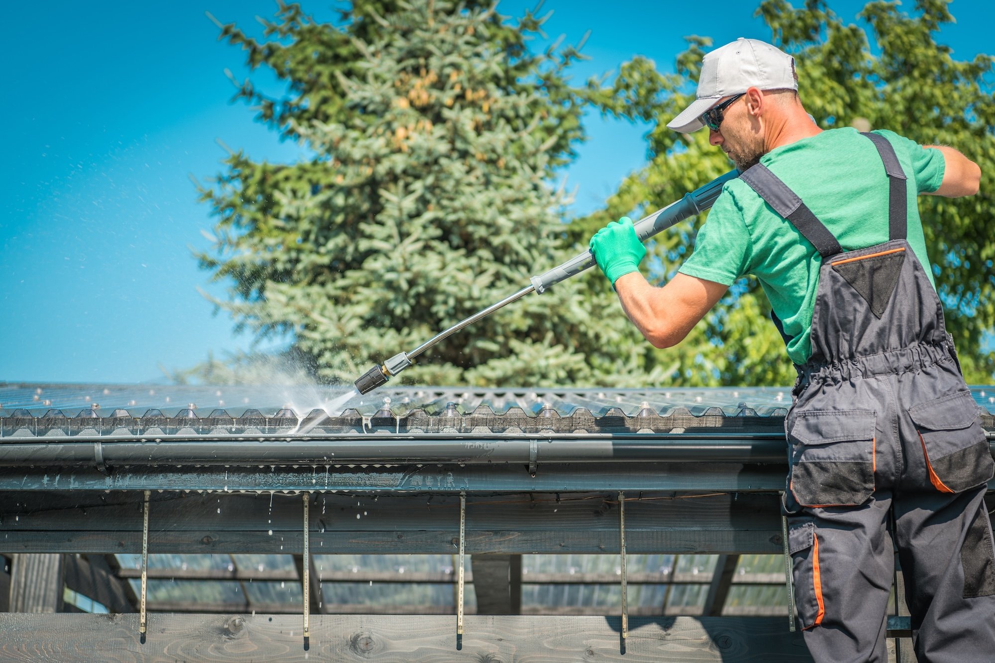 A man wearing a green shirt, gray cap, sunglasses, and gloves is using a pressure washer to clean the surface of a metal roof on a house. There are trees and a bright blue sky in the background.