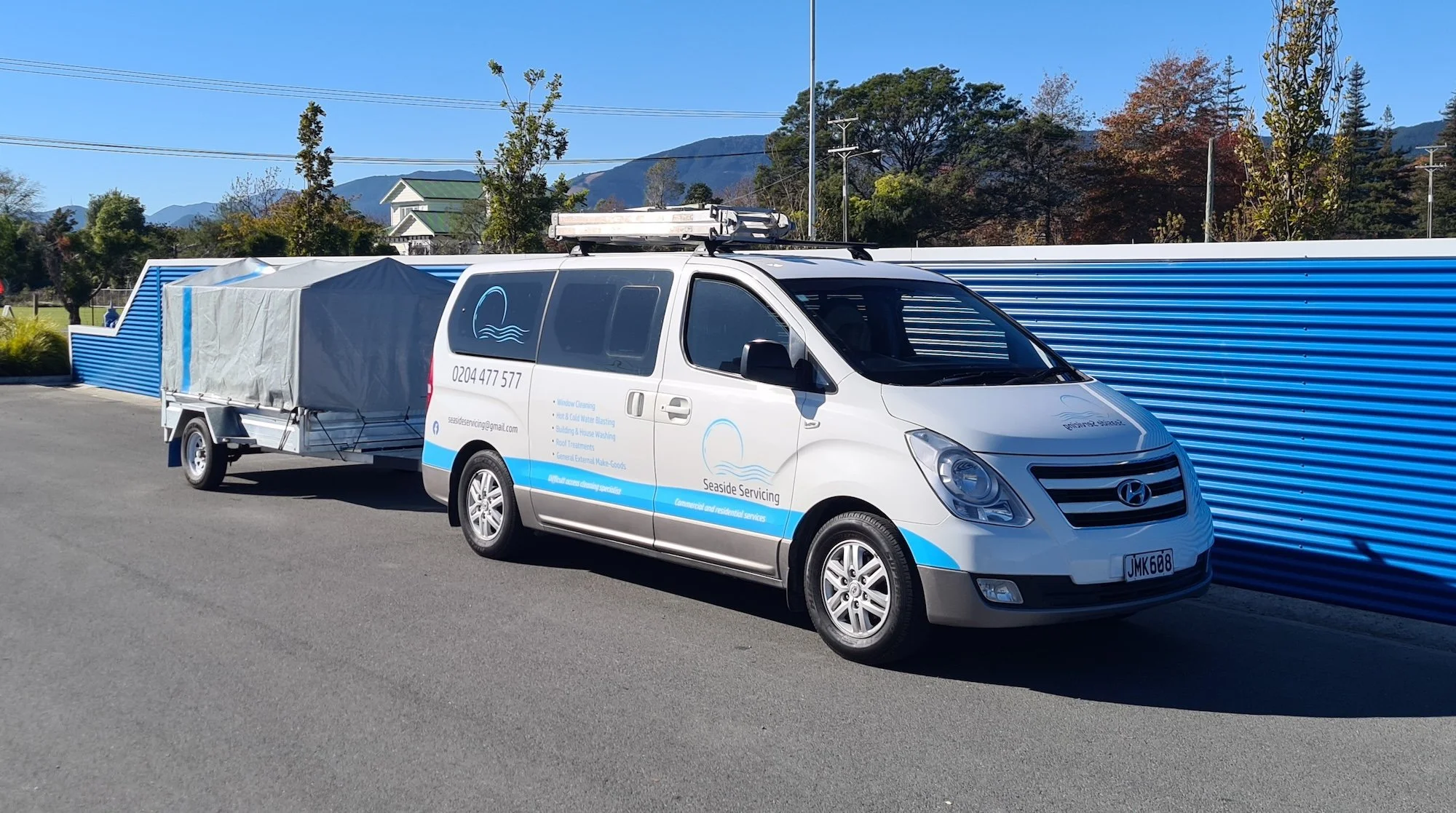 White van with blue and gray graphics parked on the street, with a trailer attached. The van has the logo and contact information for 'Seaside Servicing,' providing window cleaning and related services.