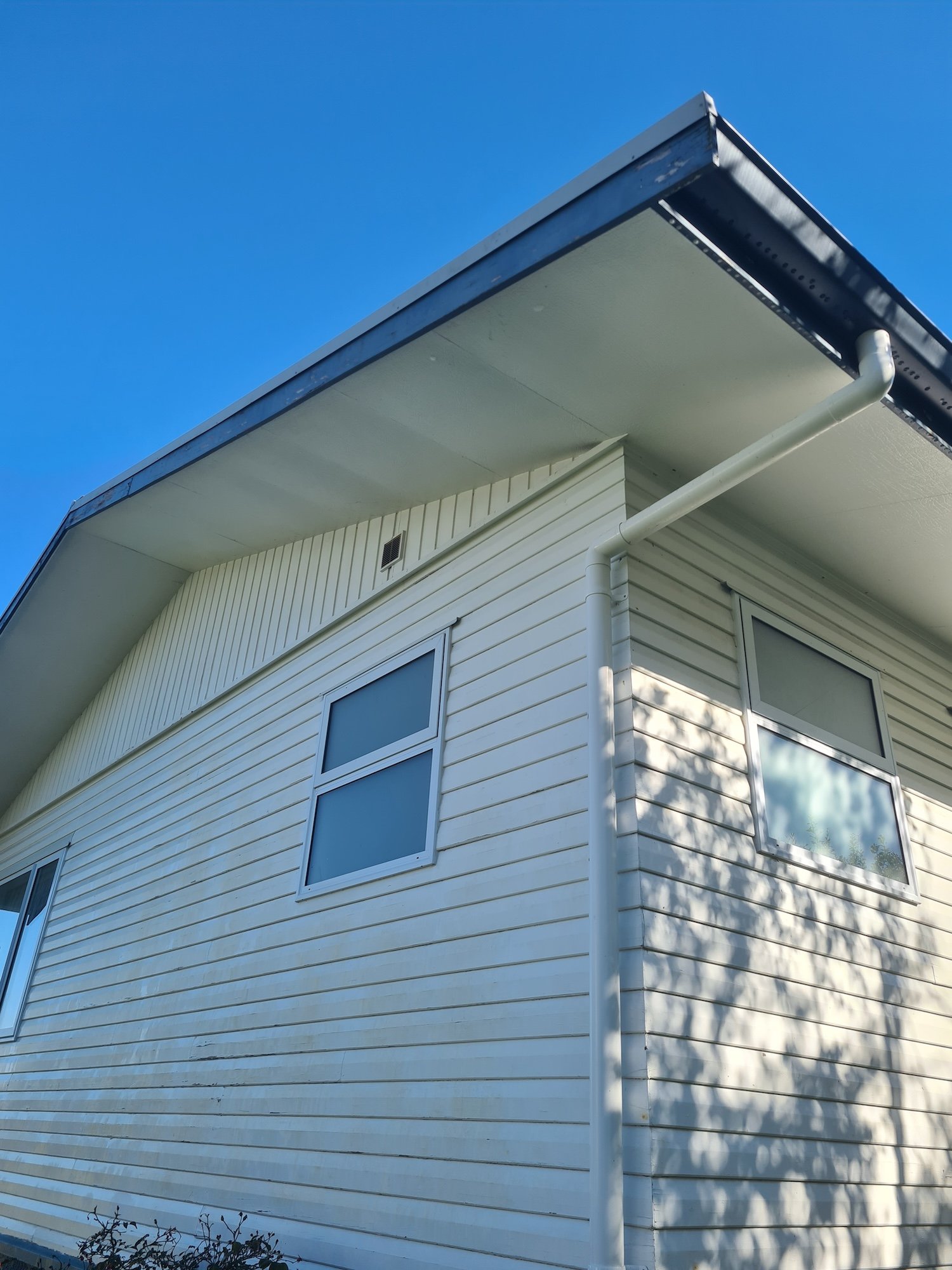 Exterior corner of a modern house with white horizontal siding, multiple windows, a white gutter, and a downspout, under a clear blue sky.