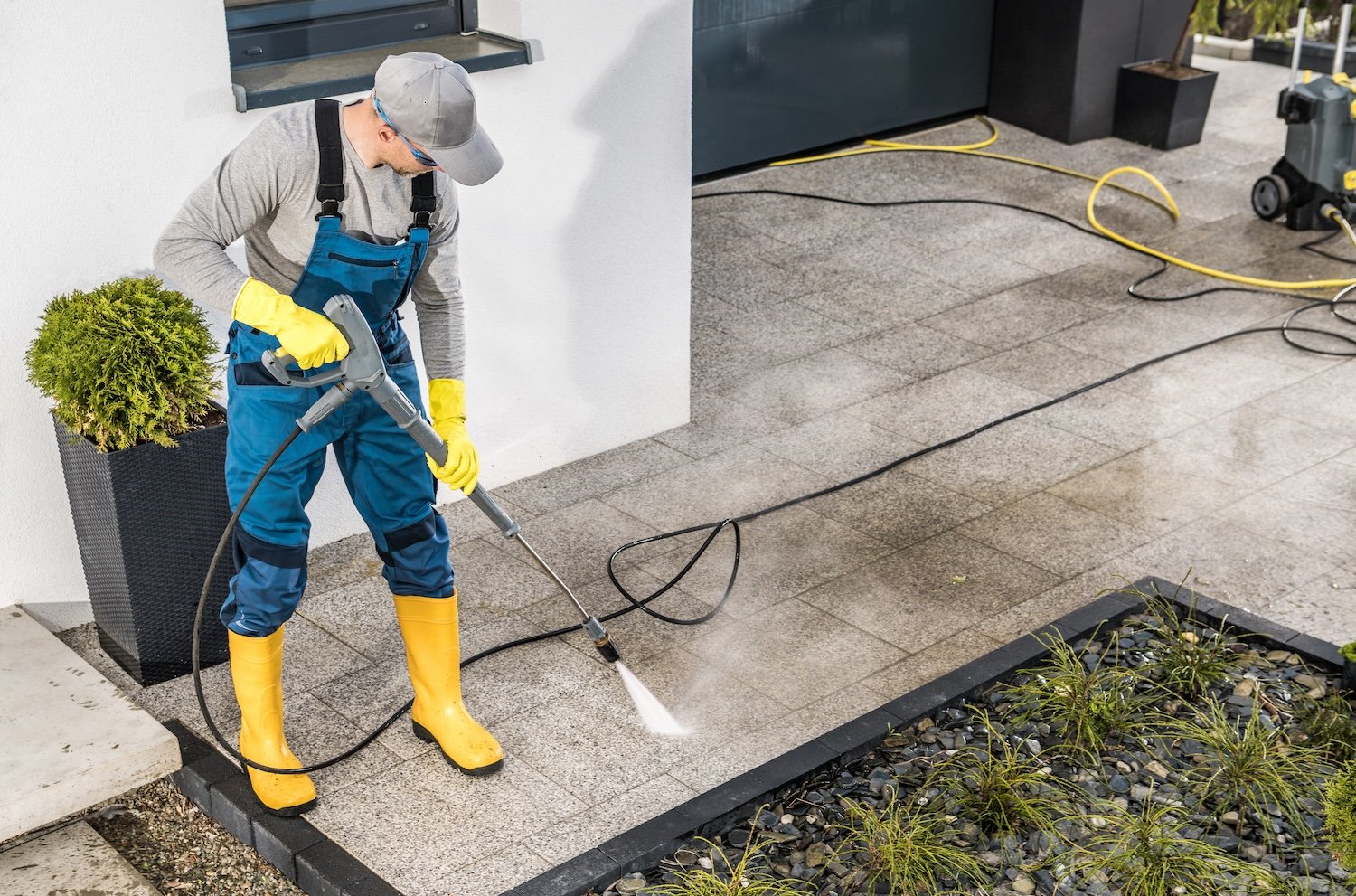 Person cleaning outdoor patio tiles with a pressure washer, wearing yellow rubber boots, gray cap, gray long sleeve shirt, blue overalls, and yellow gloves.