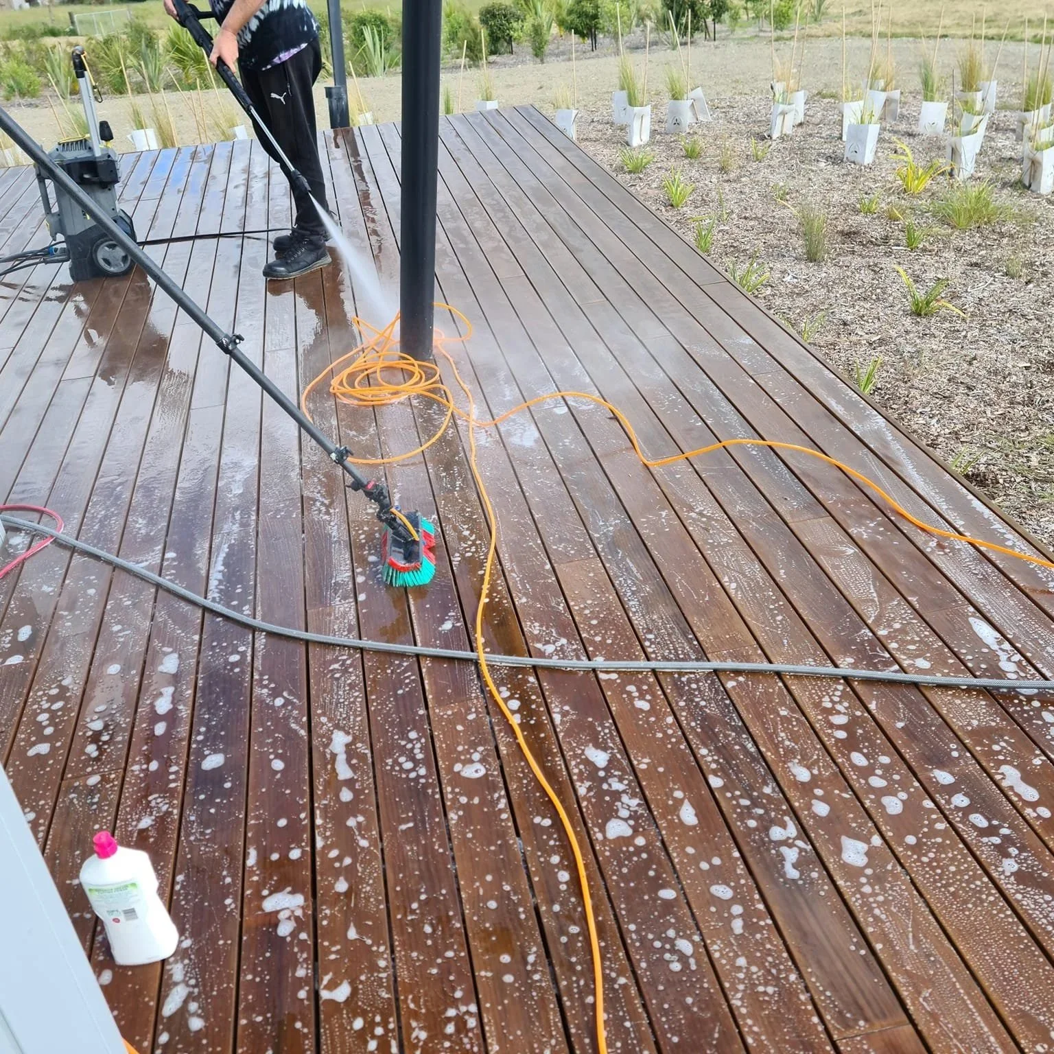A person wearing black clothes and shoes is pressure washing a wooden deck with soap, causing suds and water to spill across the surface. There are plants in the background, and cleaning equipment and an orange extension cord are visible.