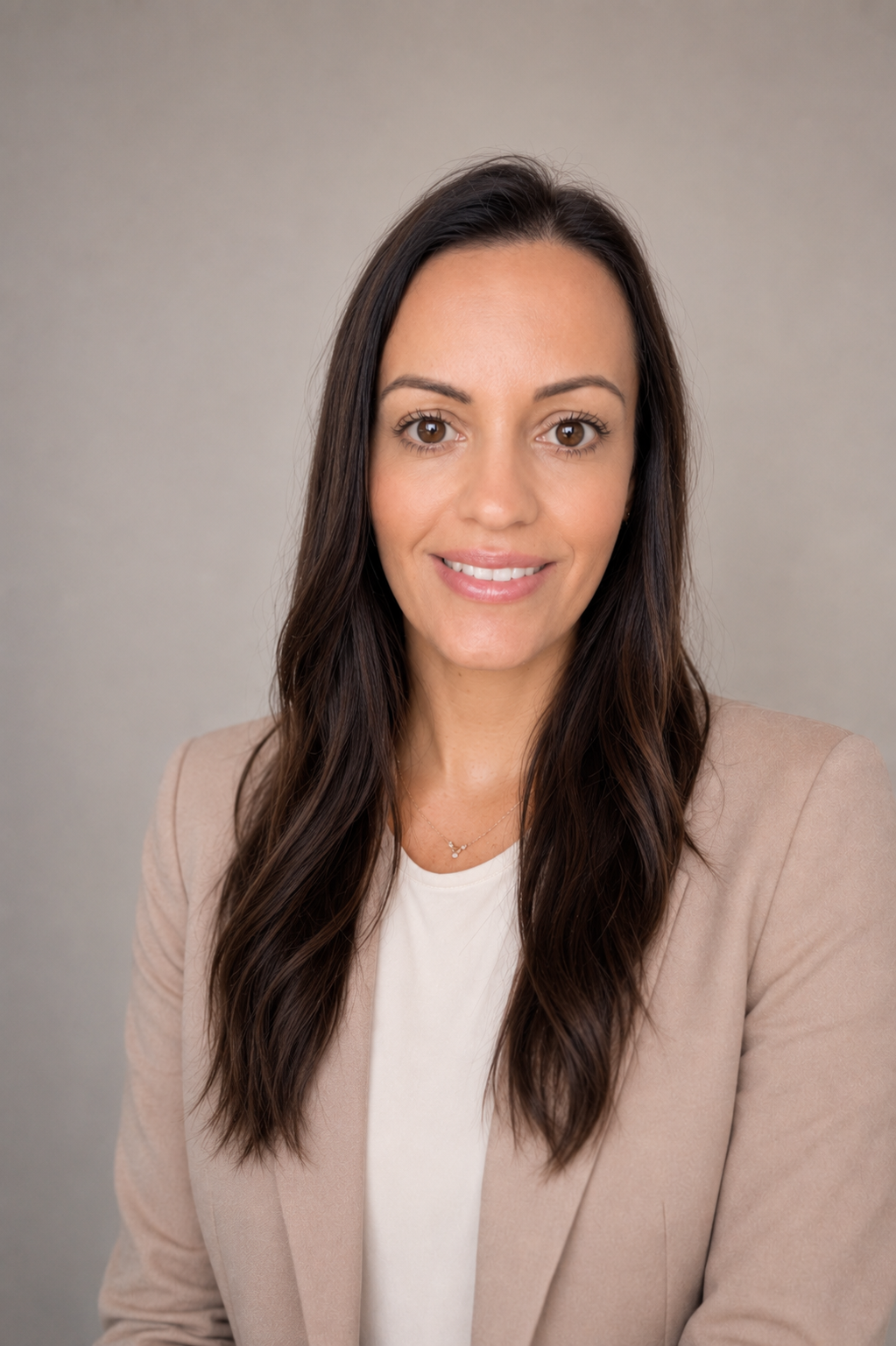 A woman with long dark hair wearing a beige blazer and white shirt, smiling at the camera against a plain gray background.