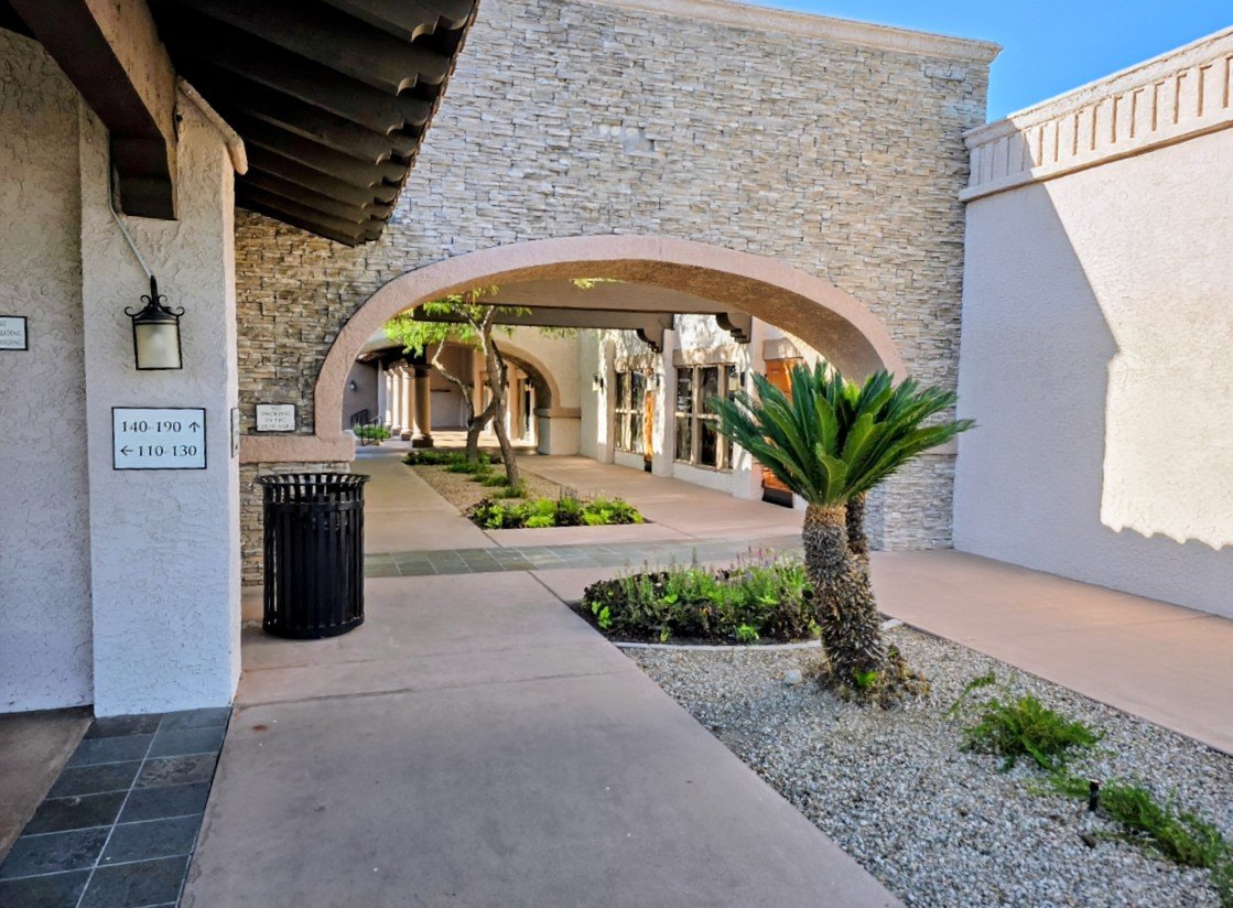 A walkway in an outdoor shopping area or mall with a stone archway, plants, and several storefronts.