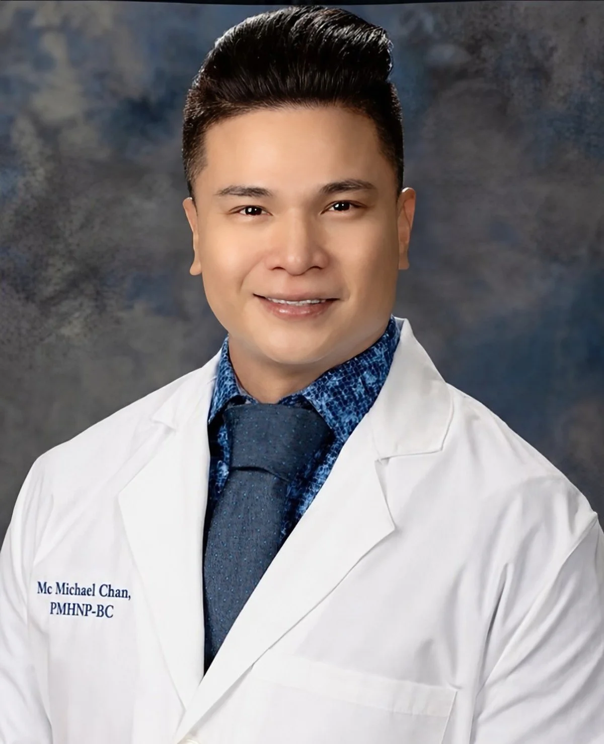 Portrait of a young man with neatly styled dark hair, wearing a white lab coat over a blue patterned shirt and a dark tie, smiling against a blurred gray background.