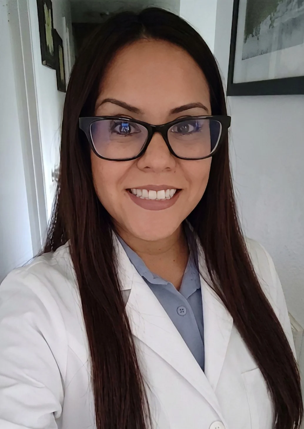 A woman wearing glasses and a white lab coat smiling for a selfie indoors.