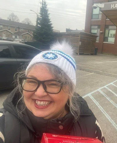 Close-up of a smiling woman wearing glasses and a knit hat with a Coldest Night of the Year emblem, outdoors in a parking lot with a car and building in the background.