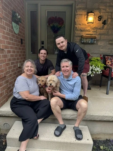 Four people and two dogs sitting and standing on a porch, smiling for a photo.