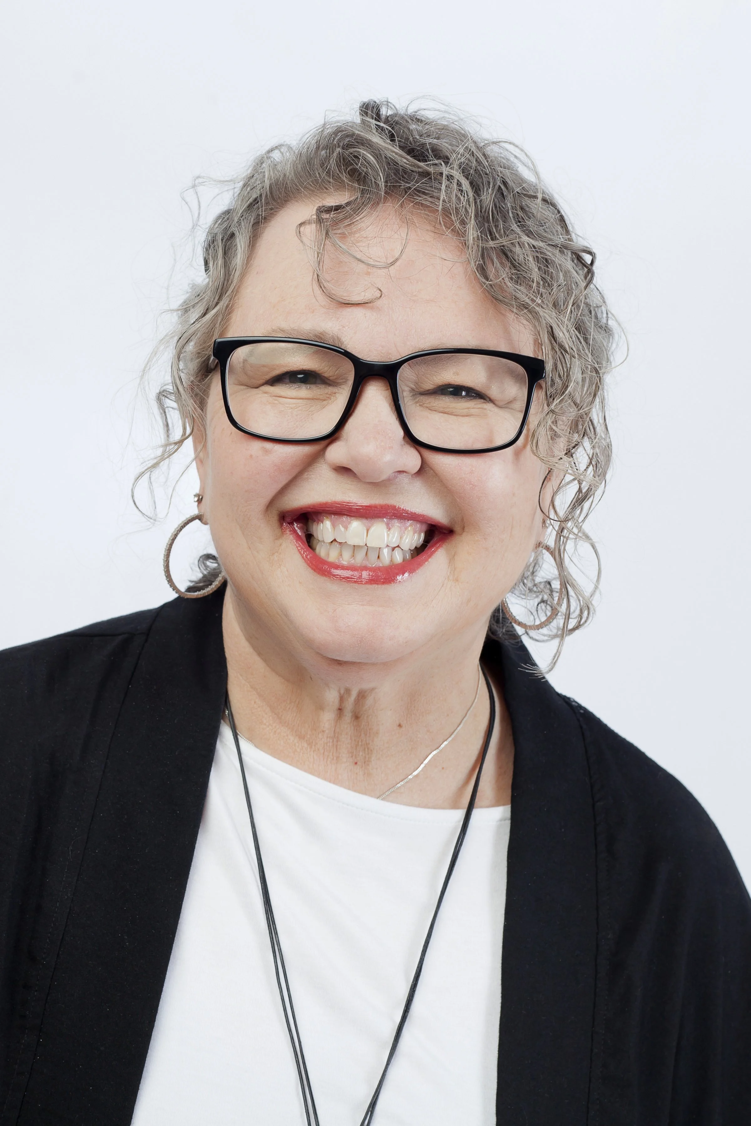 A  woman with curly hair, wearing black glasses, smiling broadly, wearing earrings, a black blazer over a white shirt, and a necklace, against a plain white background.