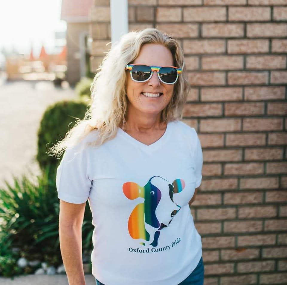 Woman with curly blonde hair wearing sunglasses and a white t-shirt with a colorful cow and rainbow design, standing outdoors near a brick wall