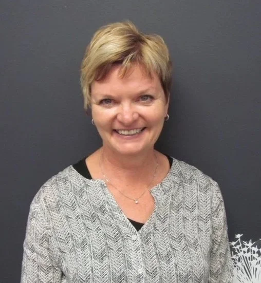 A smiling woman with short blonde hair wearing a patterned blazer over a black top, standing against a dark gray background.