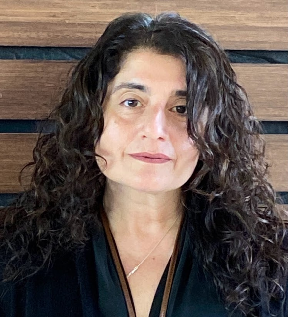 A woman with long, curly dark hair, wearing a black top and a thin necklace, sitting in front of a wooden background.