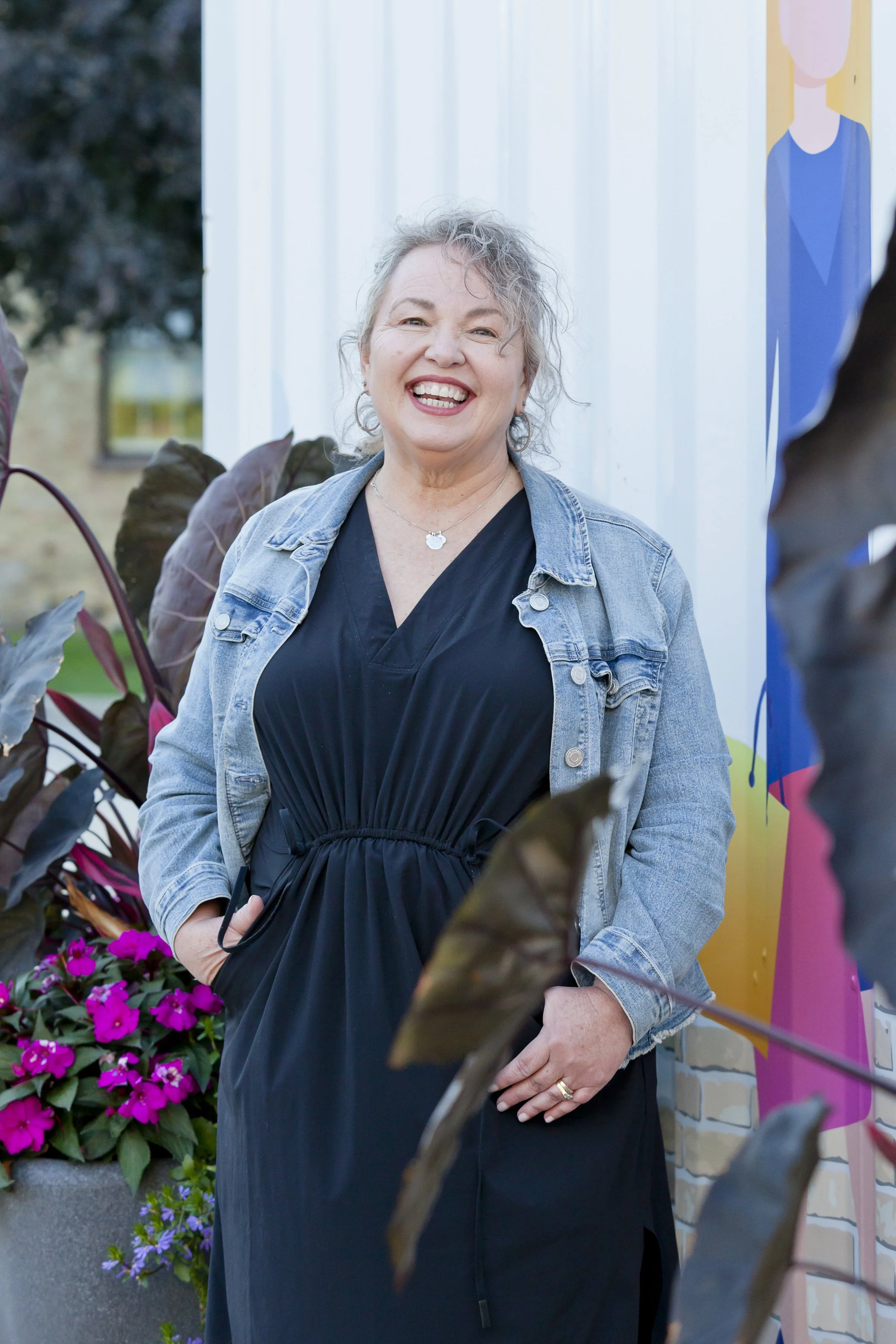 A smiling woman with short curly hair wearing a black dress and denim jacket standing outdoors near colorful flowers and a painted mural.