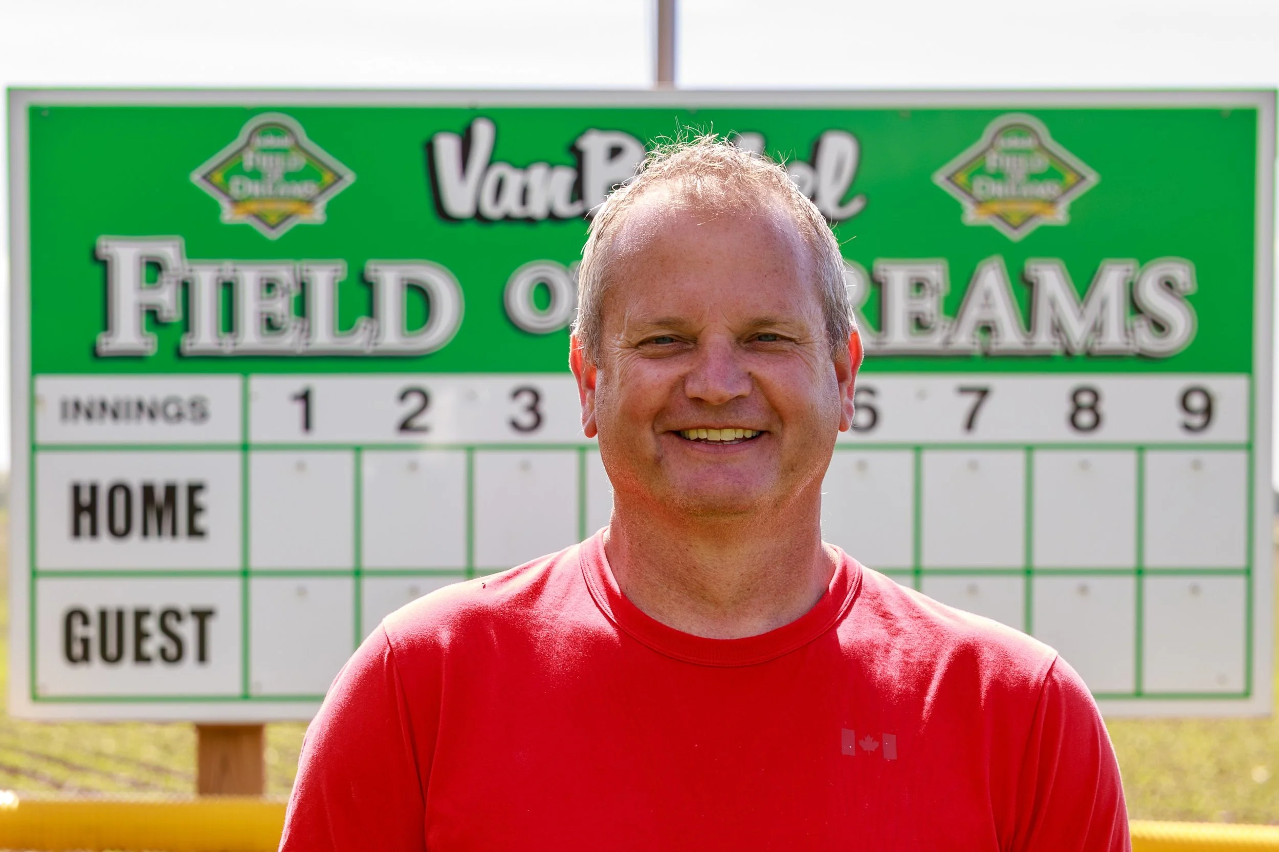 A smiling man in a red shirt standing outdoors in front of a green and white scoreboard at a baseball field.