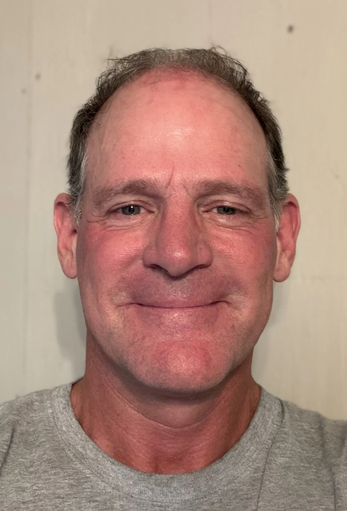 Close-up of a smiling middle-aged man with short, dark hair, wearing a gray t-shirt, standing against a plain wall background.
