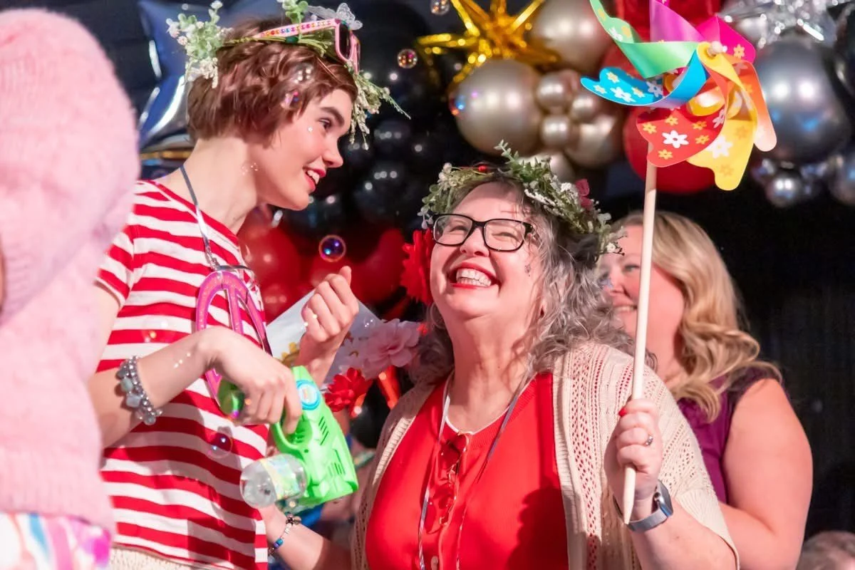 Two women celebrating at a festive event, wearing flower crowns, holding colorful pinwheels, and surrounded by balloons and decorations.