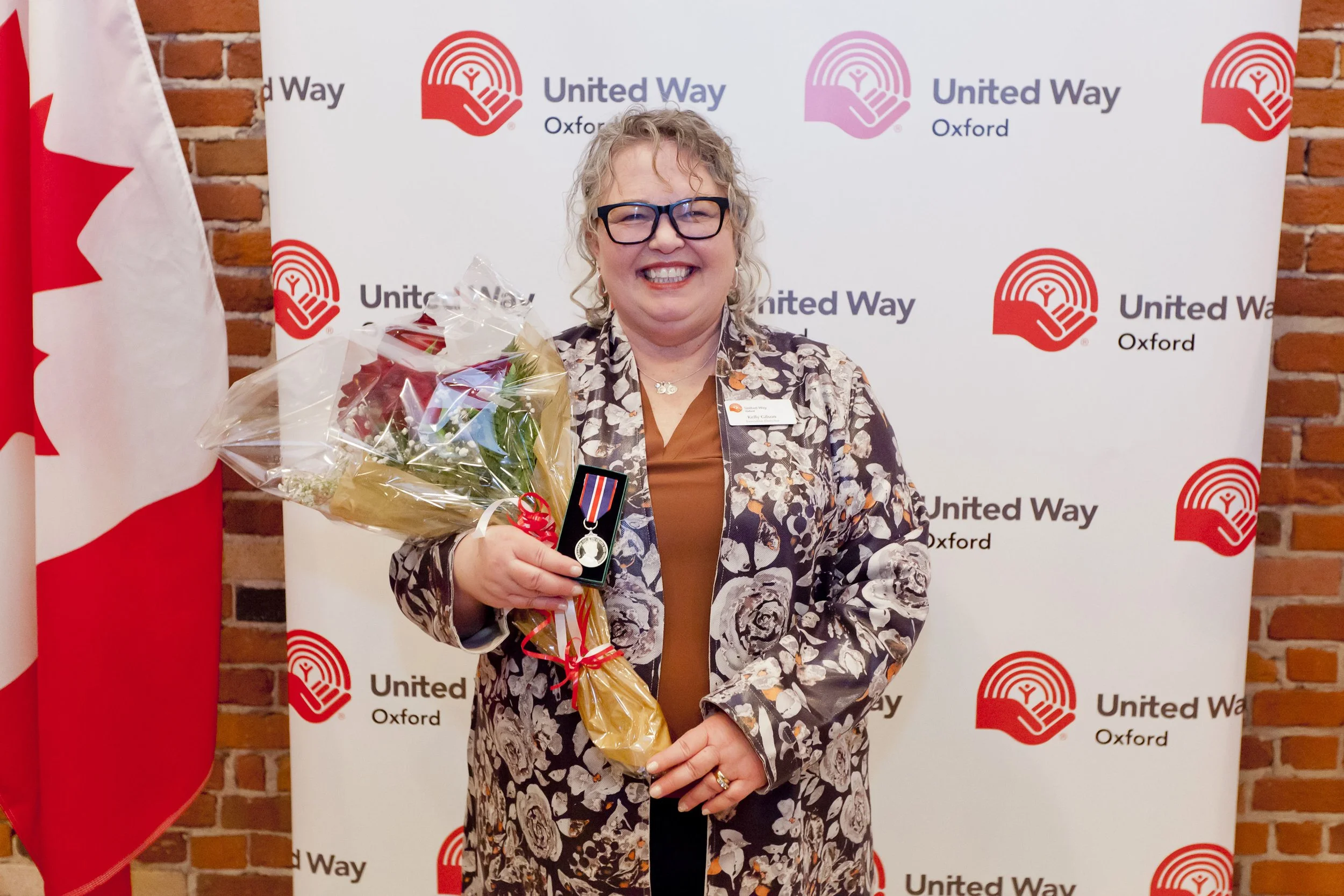 A woman holding a bouquet of flowers and a silver medal standing in front of a United Way Oxford backdrop with a Canadian flag to her left, smiling broadly.