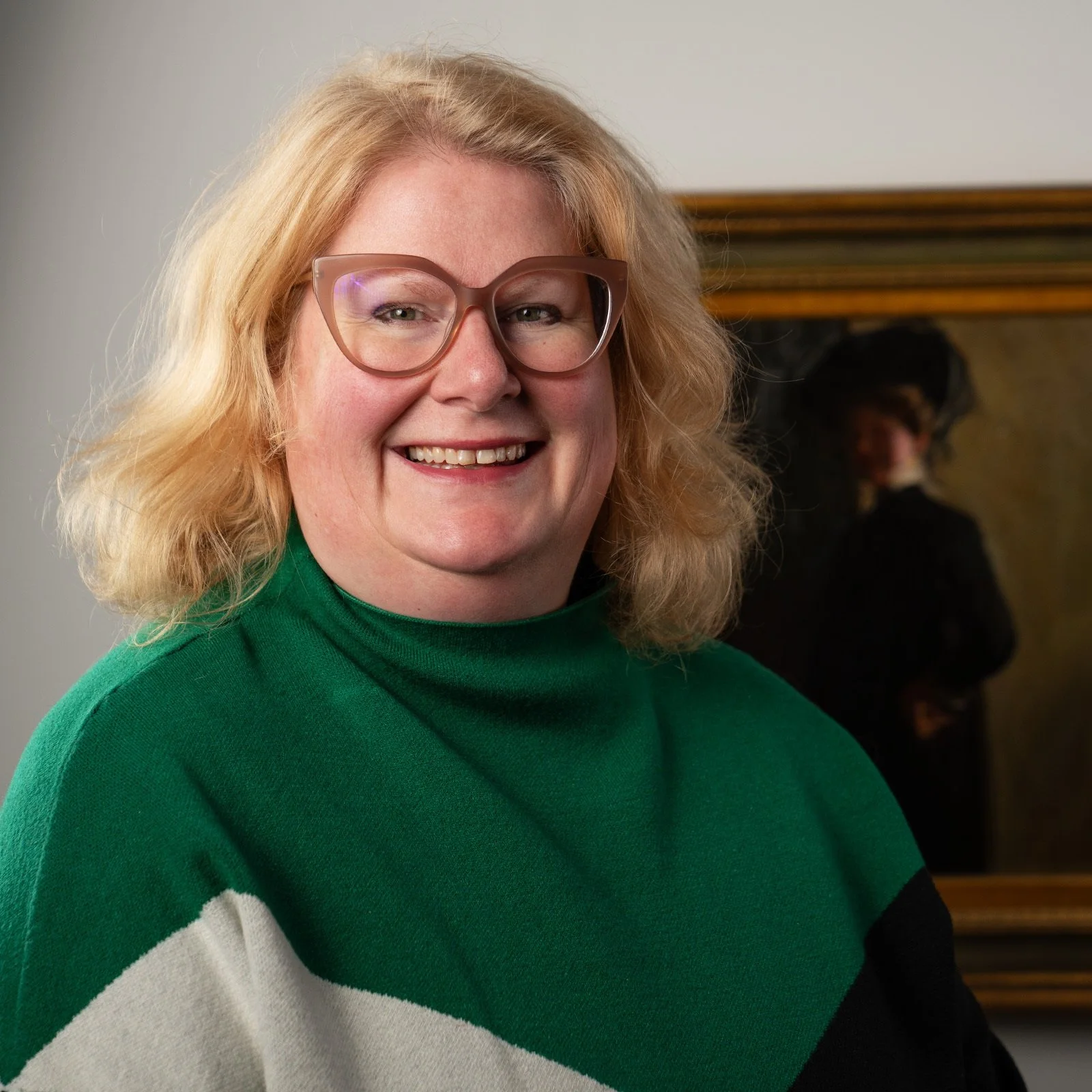 Smiling woman with blonde hair wearing glasses and a green sweater, in an indoor setting with a blurred person in the background.