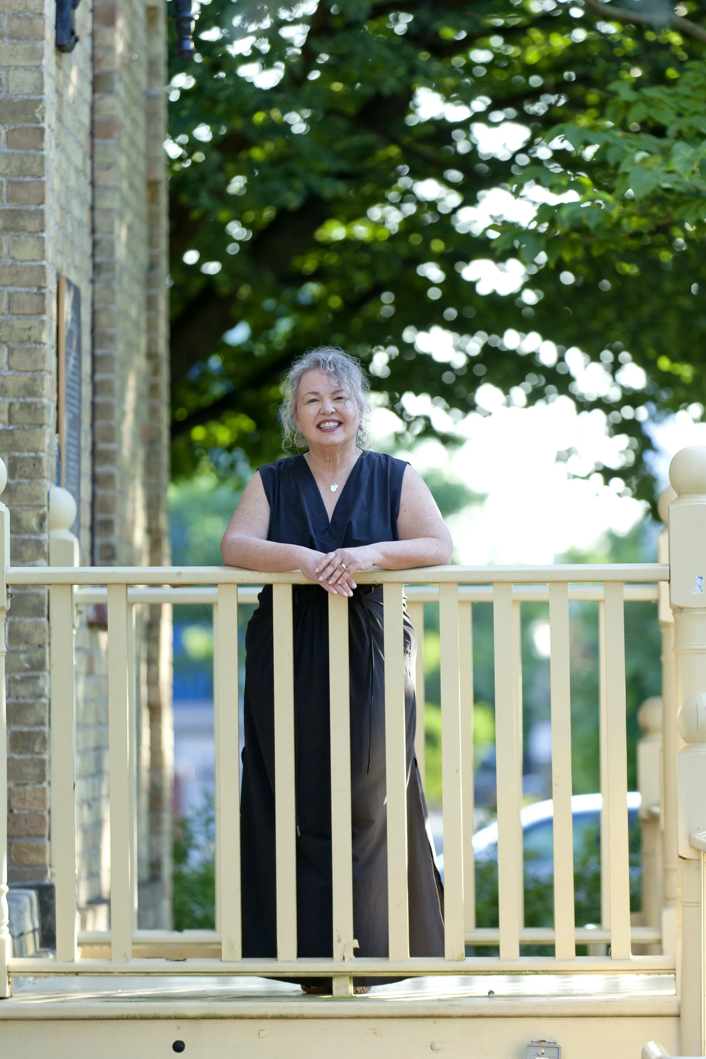 An older woman with gray curly hair smiling and leaning on a yellow porch railing outdoors, with green trees and a brick building in the background.