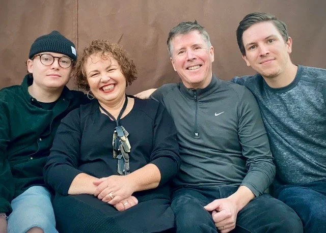 Four people sitting together on a couch, smiling, with a brown background. The group includes a mom, a dad, and two adult sons, all dressed casually.