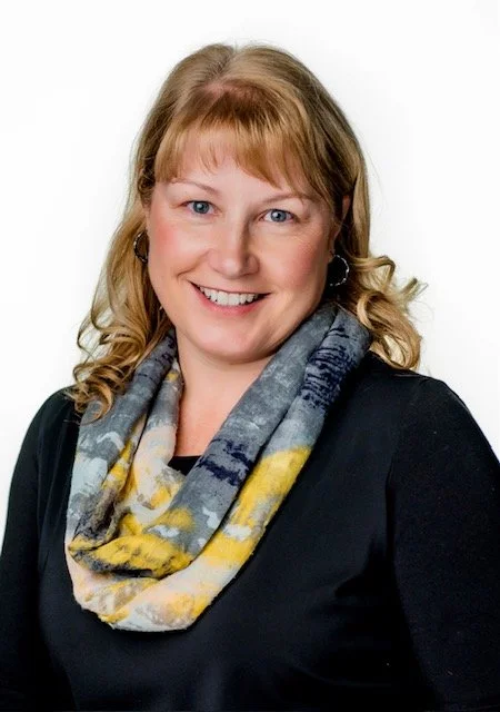 Portrait of a smiling woman with shoulder-length light brown hair wearing a black top and a colorful patterned scarf, against a white background.