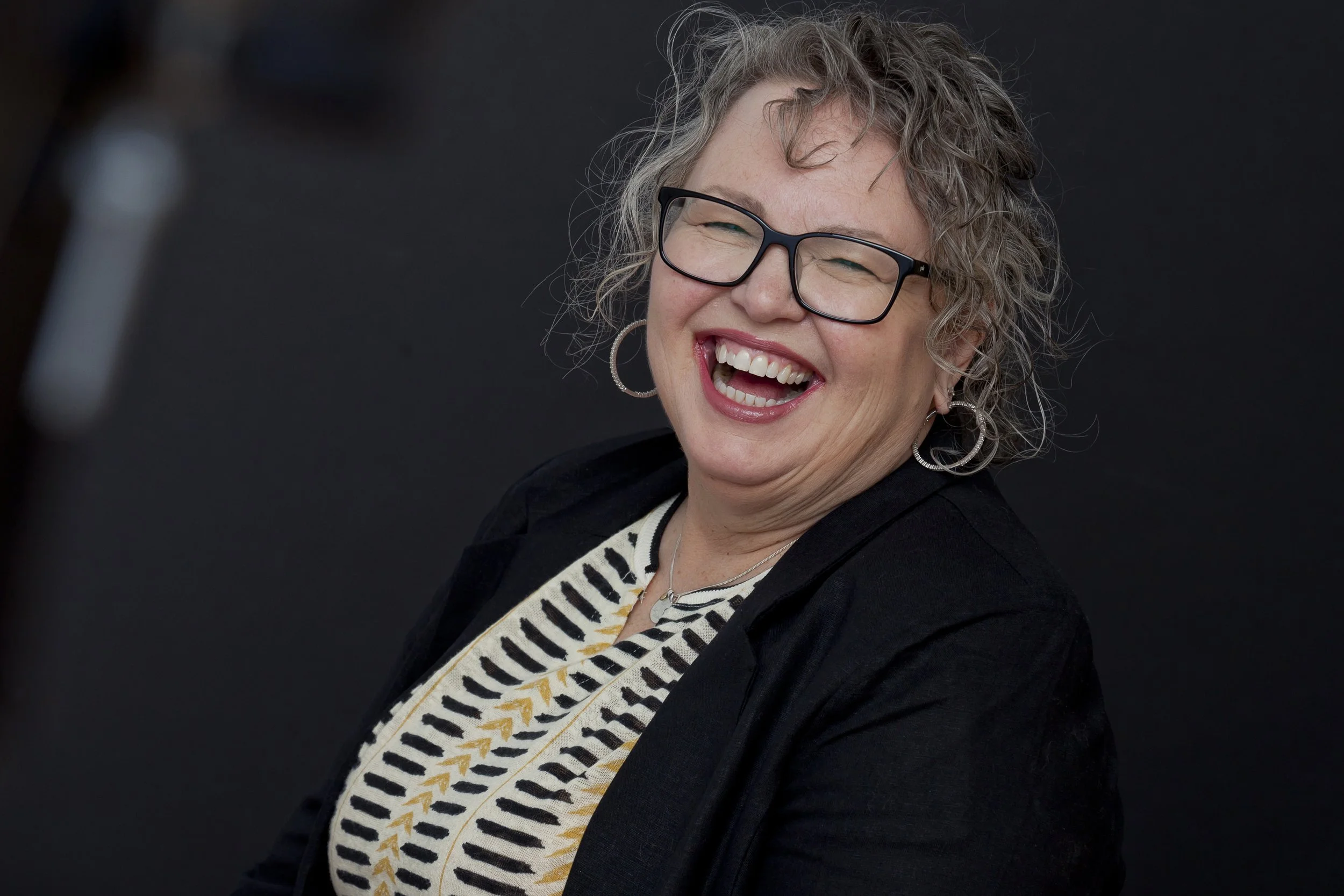 A woman with curly hair and glasses laughing, wearing a black jacket and a striped shirt with a pattern of black, white, and yellow.