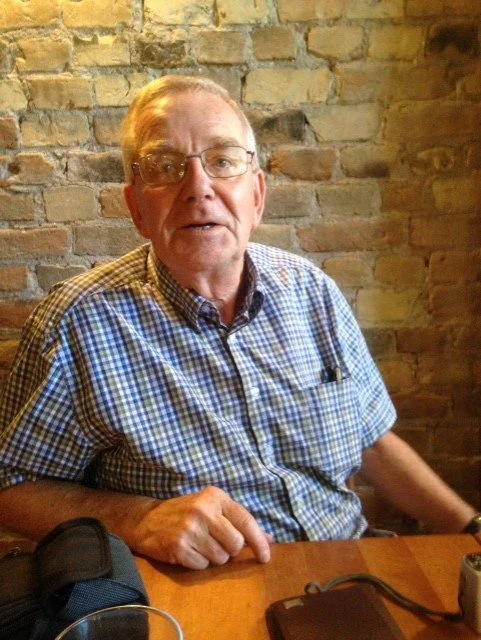 Older man with glasses wearing a blue checkered shirt sitting at a wooden table against a brick wall.