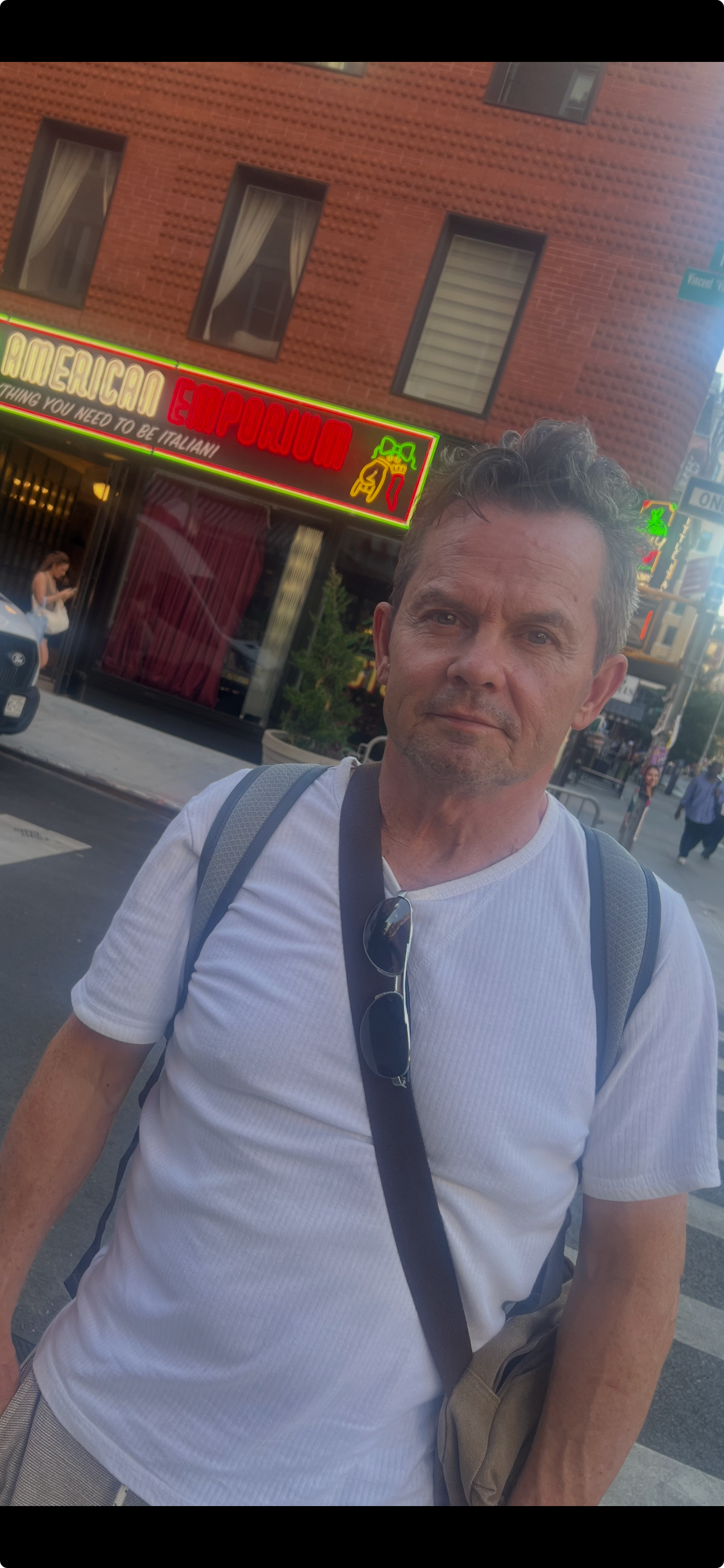 A man wearing a white shirt, standing on a city street, with neon signs and a brick building in the background.