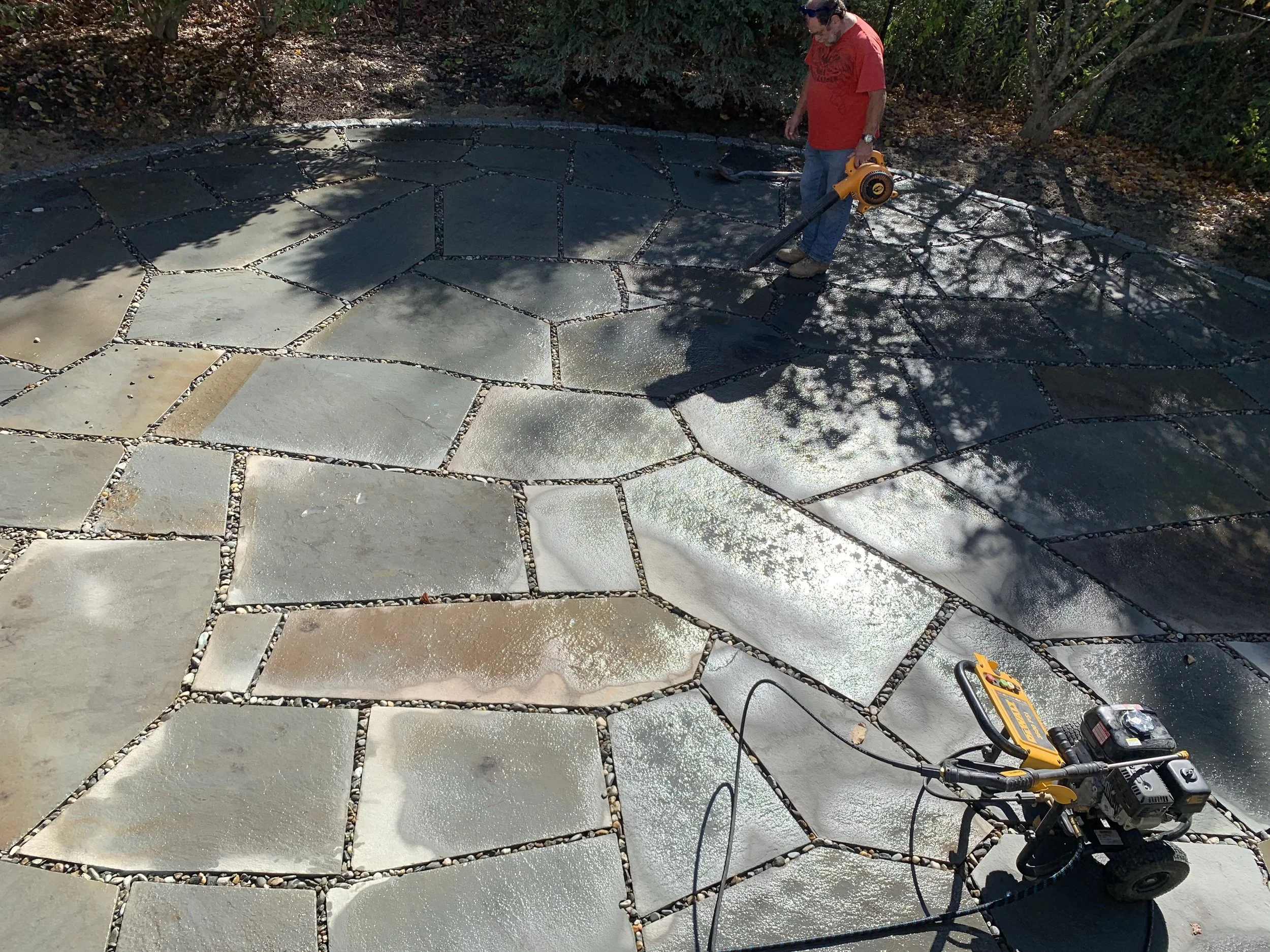 A person using a power tool to cut or grind a large stone patio with irregularly shaped stones set in mortar, in a backyard surrounded by bushes and trees.