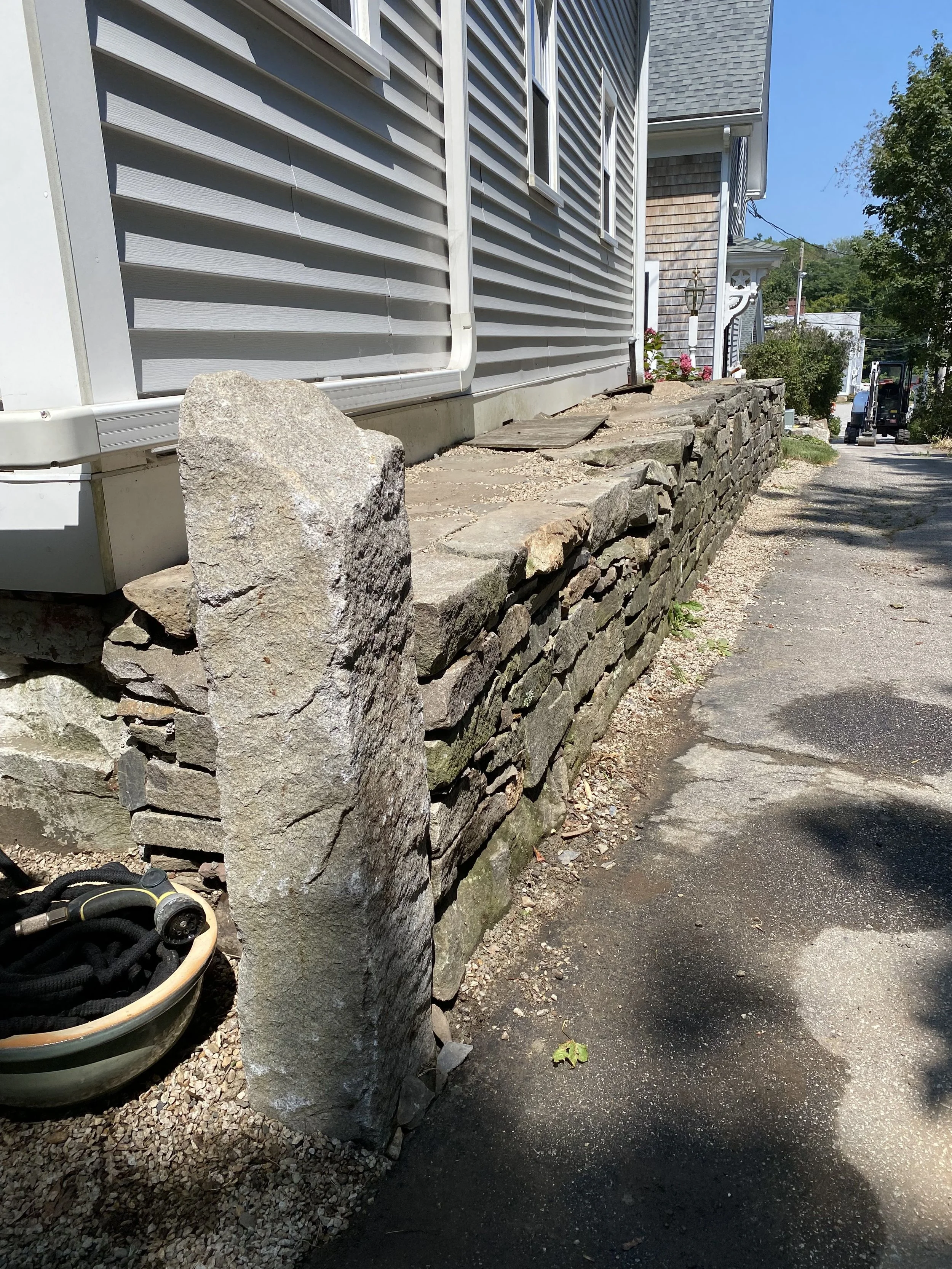 A stone retaining wall alongside a sidewalk in front of a house, with a large rock at the start of the wall, and a bucket with a hose on the sidewalk.