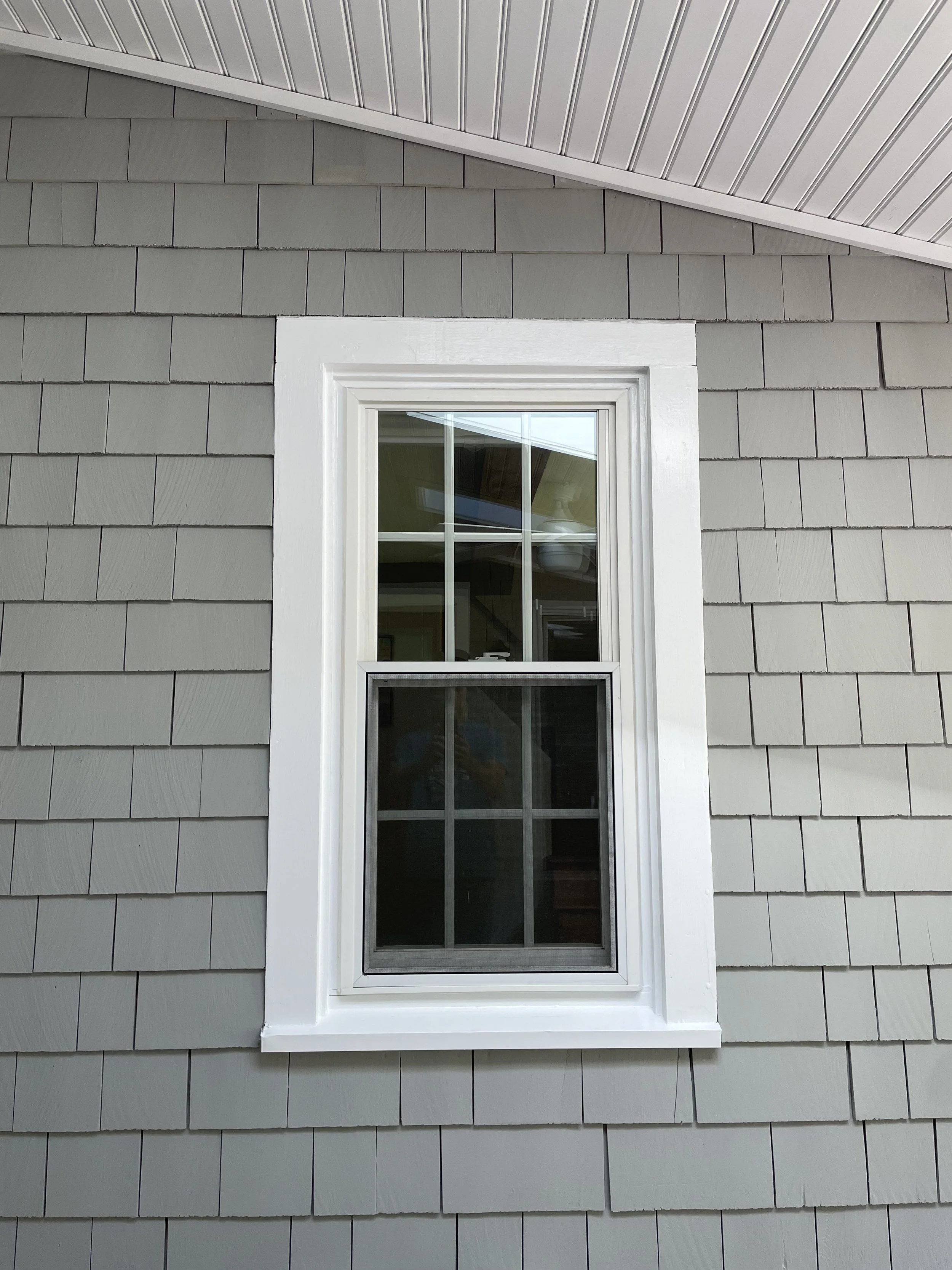 Close-up exterior window with white frame on gray shingled house wall, looking up at the roofline with gray siding and an overhanging white soffit.