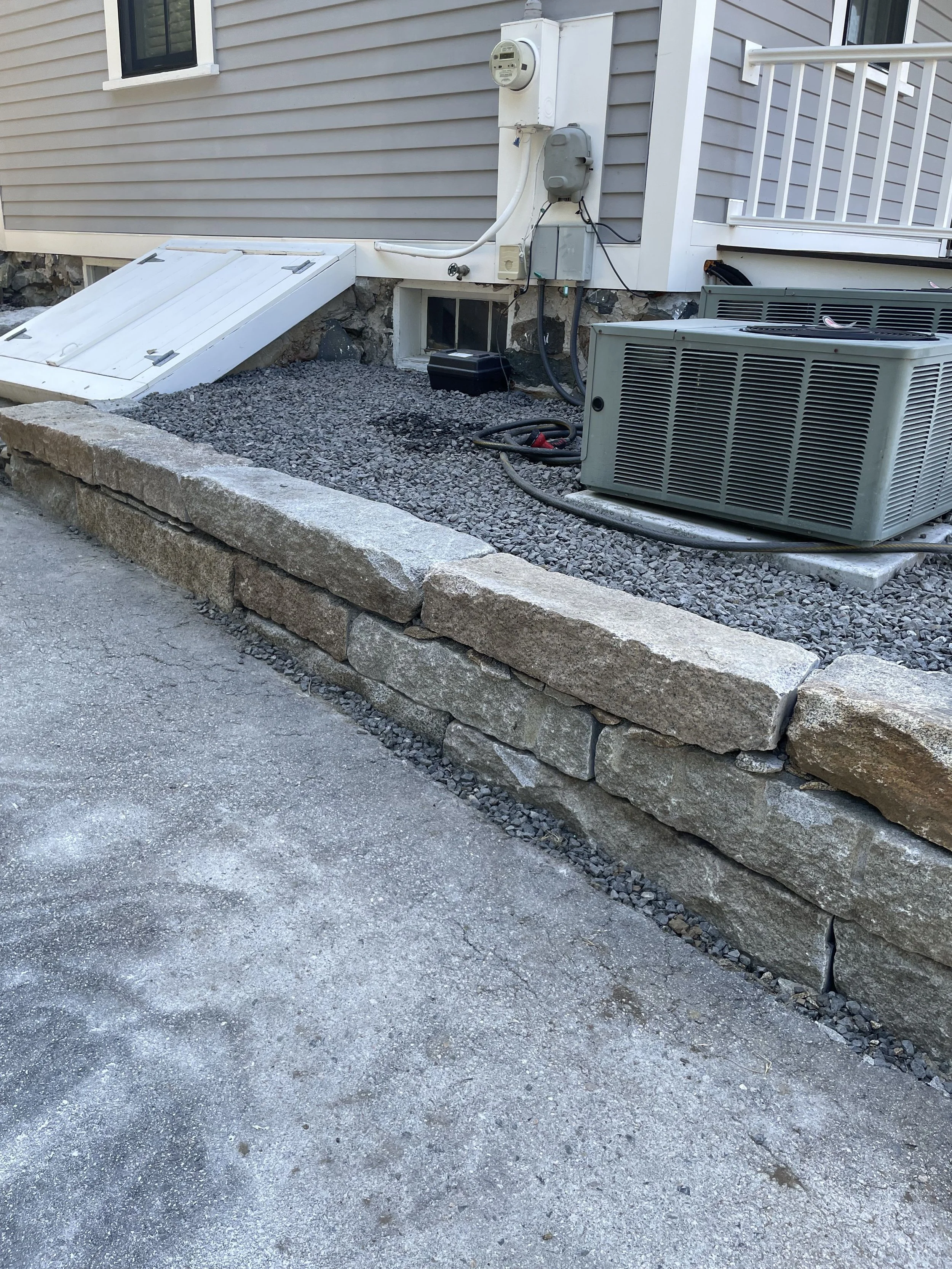 Residential house foundation with exterior fixtures, an air conditioning unit, and a gravel bed, surrounded by a stone retaining wall and a concrete walkway.