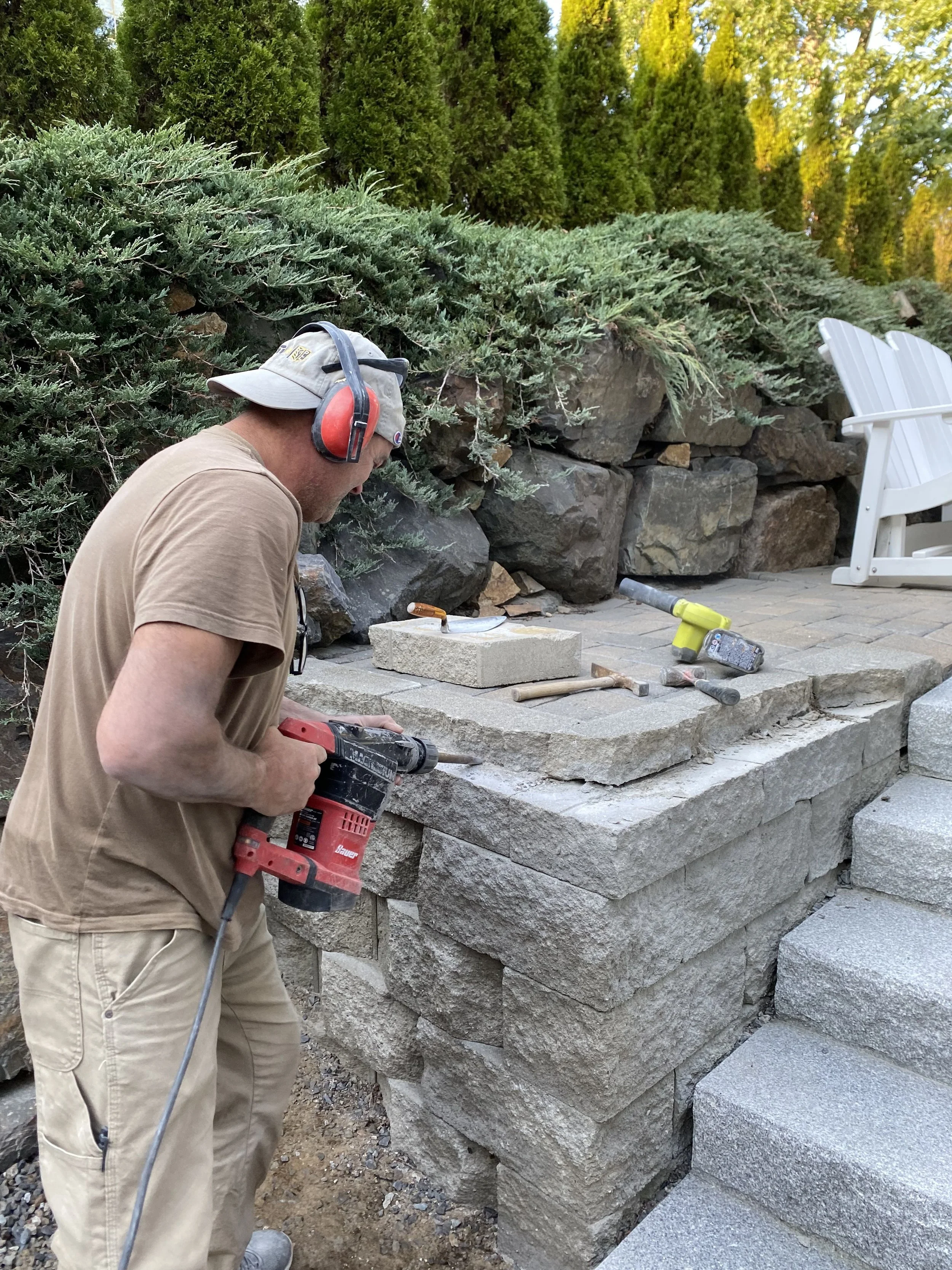 A man wearing a beige T-shirt, khaki pants, a gray cap, and red safety hearing protection is working with a cordless drill on stone steps outdoors. There are green bushes and trees in the background, with a white garden chair on the right side of the image.