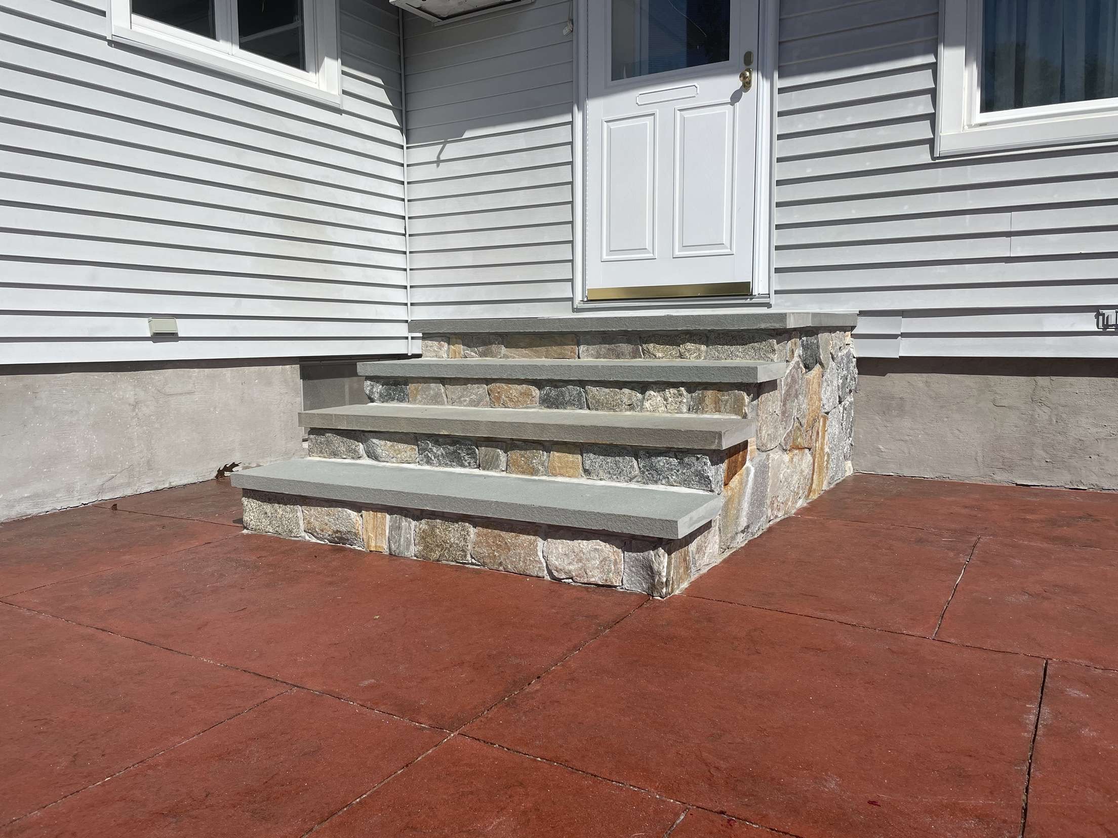 Front steps leading up to a white door with a small window, with stone and concrete foundation, light gray vinyl siding, and windows on either side.
