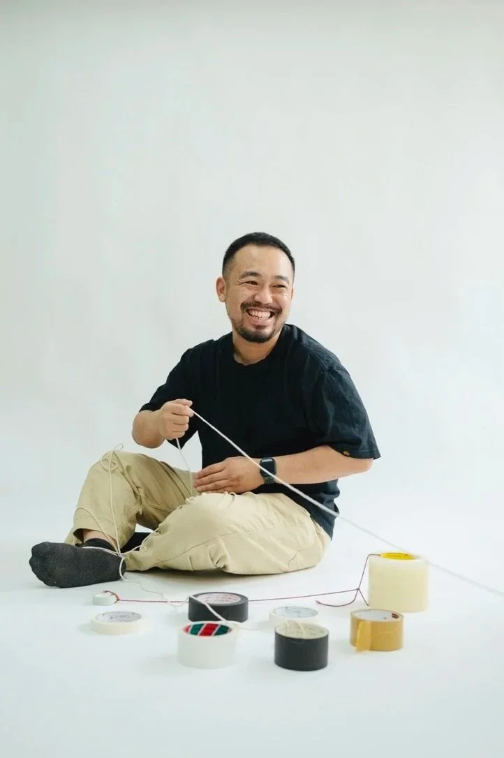 Smiling man sitting on the floor, surrounded by various rolls of colored tape, with some tape and a string in his hands.